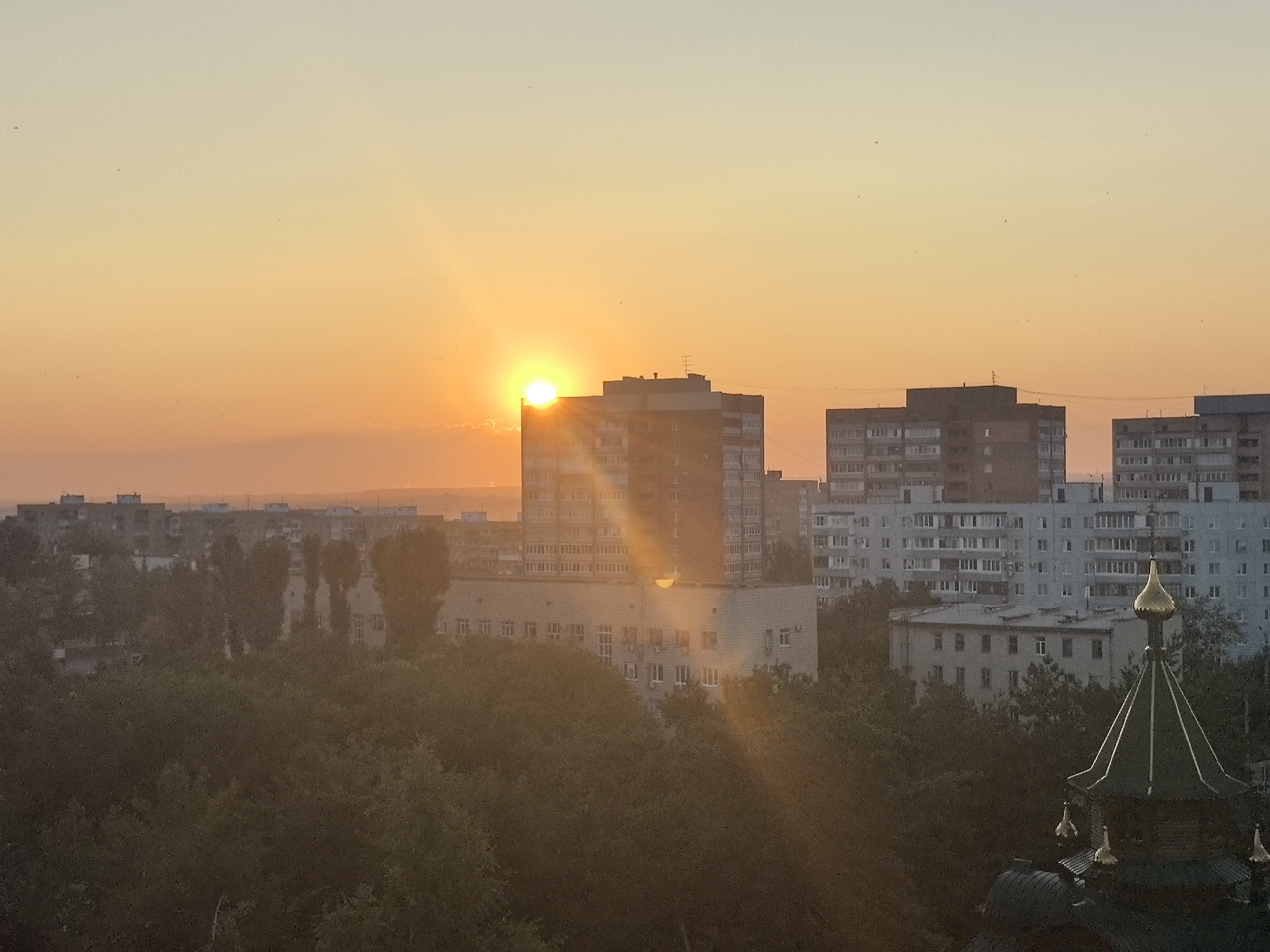 The image depicts a city skyline at sunset in Tolyatti, Russia. The sun is setting in the left portion of the frame, casting a warm orange glow across the sky and silhouetting various buildings. In the foreground, dense green trees partially obscure the lower parts of the scene. Behind the trees, several multi-story apartment buildings are visible, characteristic of Soviet-era architecture. One prominent building, a taller apartment block, is situated in the middle ground, with the sun partially behind it, creating a lens flare effect. To the right, the ornate golden dome of a church or religious building peeks out from behind the trees, its intricate design a stark contrast to the utilitarian apartment blocks. The sky is a gradient of soft orange and yellow, with a few faint clouds visible near the horizon. Small dark specks, possibly birds, are scattered across the sky. The overall impression is one of urban landscape bathed in the gentle light of dusk.