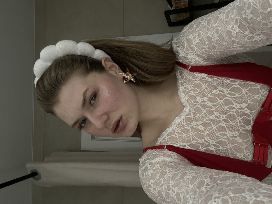 A woman with light skin, light brown hair in a ponytail, and blue eyes looks into the camera. She wears a white, puffy headband and star-shaped earrings. She is wearing a white lace top with a red harness. The background includes a wall, a shelf with objects on it, and a window with a beige curtain. The image is rotated, with the subject appearing at an angle.