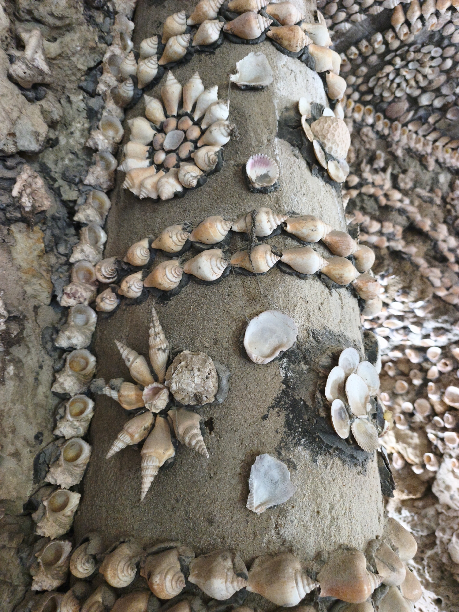 The image depicts a textured, rough concrete column densely adorned with various seashells. These seashells are meticulously arranged in patterns and clusters, forming decorative elements on the column's surface. Some shells are whole and pointed, resembling conch shells, while others are smaller, spiral-shaped, or appear as fragments. The shells exhibit a range of natural colors, predominantly in shades of white, cream, beige, and light brown, with some showing subtle pink or grey hues. The concrete surface itself has a coarse texture and is varied in color, with patches of darker grey or black interspersed with lighter sandy tones. The background is filled with more clusters and arrangements of seashells, suggesting that this column is part of a larger structure or environment extensively decorated with them. The overall scene is static and focuses on the intricate decorative details of the seashell-encrusted column. This structure is located in the Tu Van Pagoda, also known as the Nui Mot Pagoda, in Cam Ranh City, Khanh Hoa Province, Vietnam.