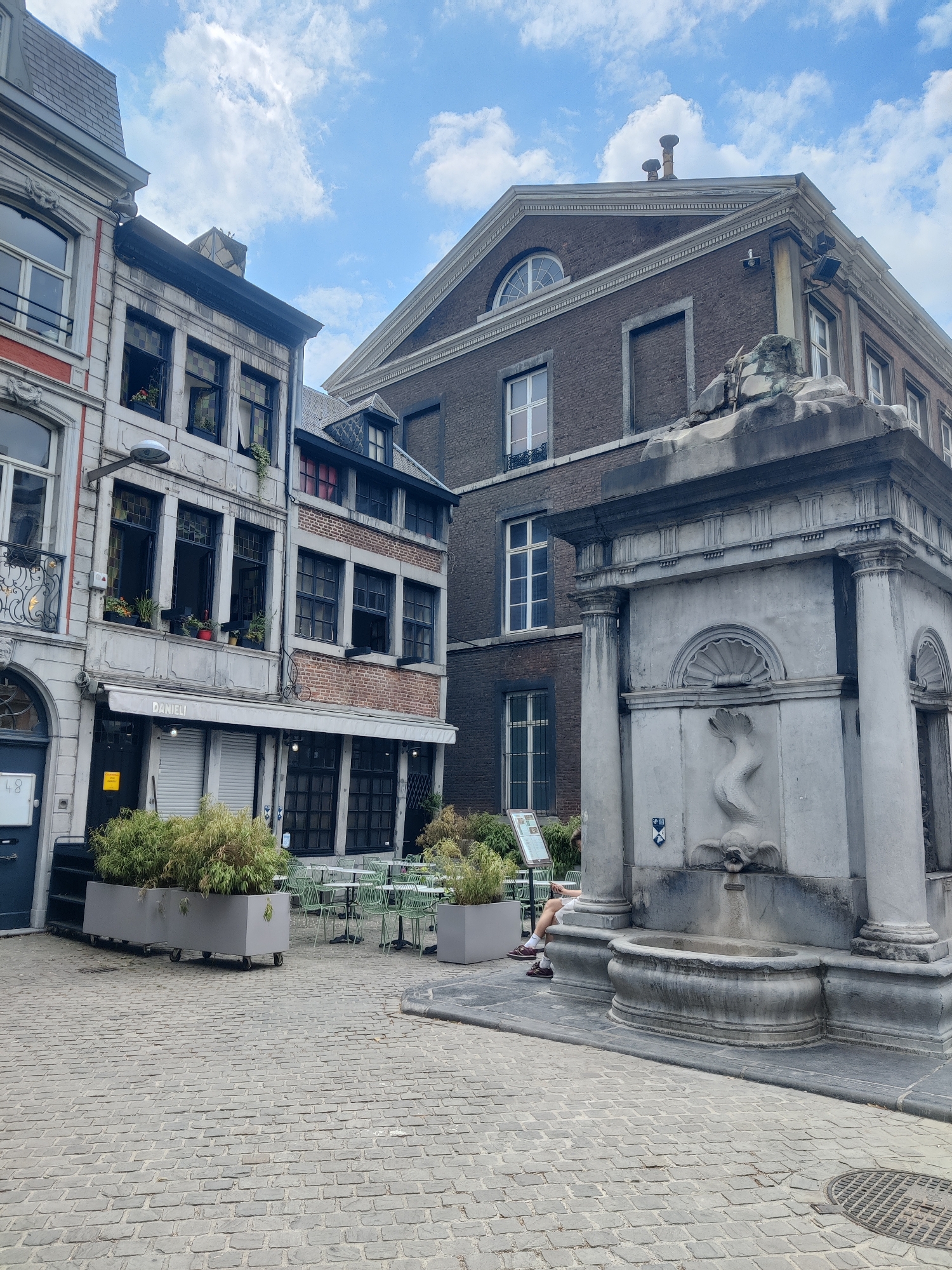 The image depicts a cobblestone square in Liège, Belgium, featuring historic architecture and a stone fountain. In the foreground, a large, ornate stone fountain with a dolphin-like spout is prominent. To the left of the fountain, a multi-story building with a red brick facade and multiple windows stands. A shopfront with a white awning labeled "DANIELI" is visible on the ground floor. Planters filled with lush greenery are placed in front of the shop and extending towards an outdoor seating area with small, round tables and green chairs. In the background, another building with a dark brick facade and a triangular pediment can be seen. The sky is partly cloudy, with patches of blue sky and white clouds. A person is seated on a low wall to the right of the fountain, partially obscured by the fountain's structure. The overall scene conveys a sense of urban tranquility and historical charm.