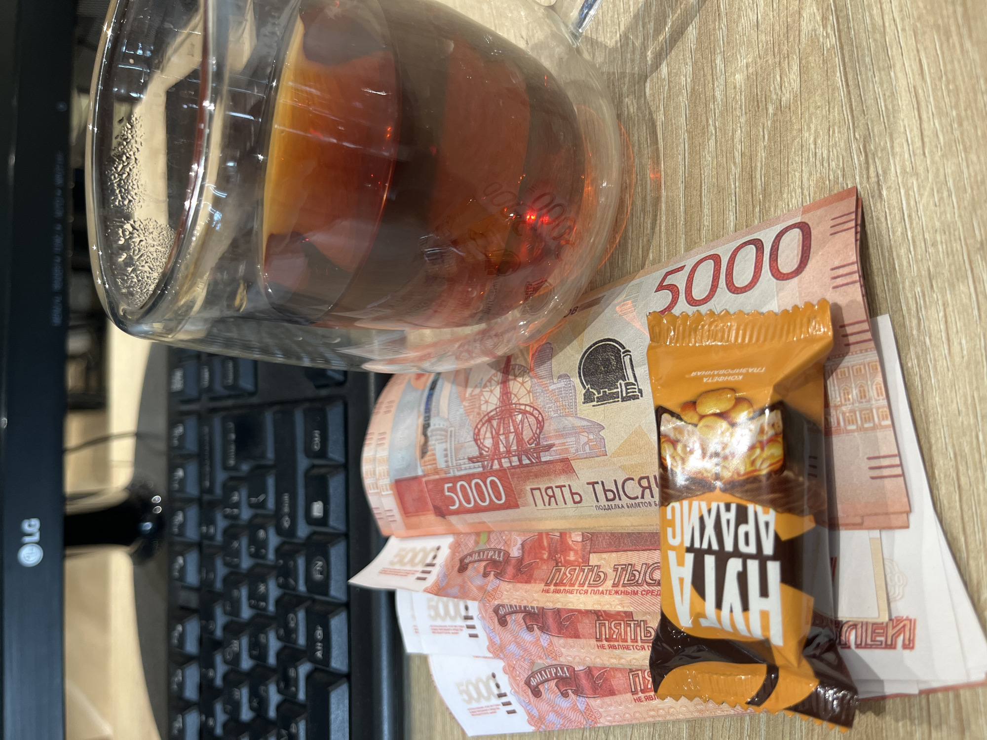 On a light brown wooden surface, partially covered by a black computer keyboard on the left and an LG monitor in the background, is a collection of objects. A clear glass mug filled with a dark amber liquid, possibly tea, sits near a stack of five red and white 5000 Russian Ruble banknotes. Resting atop the banknotes is a small, orange and brown candy bar wrapper with Cyrillic writing, displaying the words "АРАХИС НУГА." The scene appears to be set on a desk, and the objects suggest a break or a financial transaction.