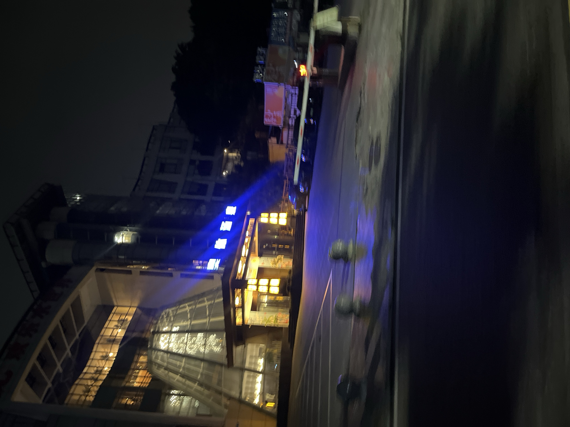 The image shows a blurry nighttime scene in front of a modern building with a glass facade and illuminated windows. The building entrance is covered by an awning with warm lighting. To the right, a truck is parked near a gate. The foreground features a dark, wet surface, likely a road or parking lot, with some bollards. The overall atmosphere is dark and somewhat indistinct due to motion blur. The scene is located in Jiulongpo District, China.