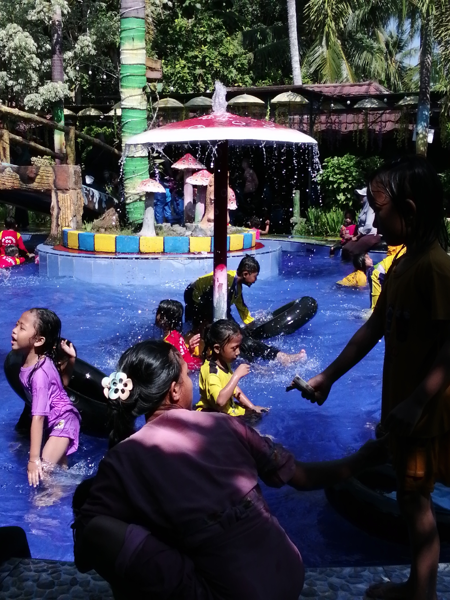 This image captures a lively scene at a water park in Tegalwangi, Indonesia, likely on a sunny day. The foreground is dominated by the pool's bright blue water, with several children actively engaged in water play. A young girl in a purple swimsuit is visible on the left, splashing in the water, with an adult figure in a pink top behind her. To the right, another child in a yellow shirt is being assisted by an adult.
In the middle ground, more children are enjoying the water. One boy in a yellow and black outfit is holding onto a large black inner tube, while another child in yellow floats on their back. Further back, a fountain feature with a red and white umbrella-shaped structure sprays water, and mushroom-shaped decorative elements are visible. The background reveals lush green foliage and palm trees, typical of the tropical climate. Hints of wooden structures and decorative elements, such as the colorful base of the fountain and a wooden railing, suggest a well-developed recreational area. The presence of multiple individuals and the overall atmosphere indicate a popular spot for family outings.