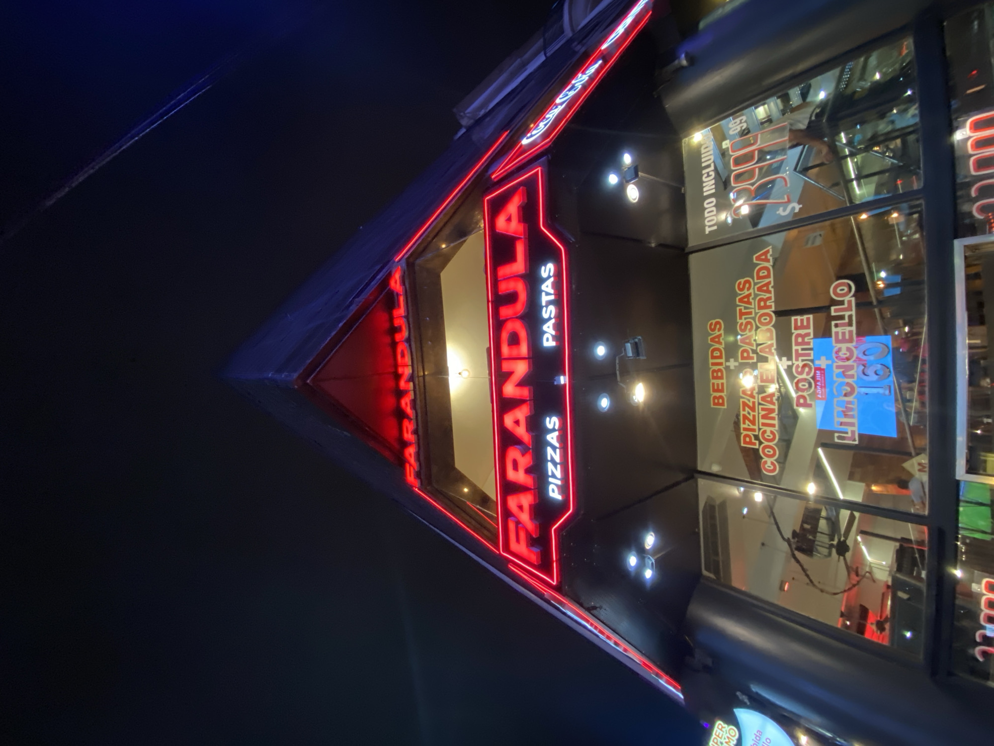 The image depicts the exterior of "FARANDULA," a pizza and pasta restaurant, at night, located at Corrientes 2450 in the city of Buenos Aires, Argentina. The building features a distinctive modern, upward-sloping triangular facade, primarily clad in dark material. Bright red neon signs illuminate the restaurant's name, "FARANDULA," displayed prominently across the upper section, with "PIZZAS" and "PASTAS" indicated below. Additional red neon trim outlines architectural elements.
Below the main signage, multiple windows provide views into the illuminated interior. Through the glass, various internal signs are visible, including "BEBIDAS," "PIZZAS PASTAS COCINA ELABORADA," "POSTRE LIMONCELLO" with a price of "160" (presumably Argentine Pesos), and "TODO INCLUIDO" (All Inclusive) with a price of "$2399.99". A ceiling fan is discernible through one window. A person in a light-colored shirt is vaguely visible leaning against an interior railing in a right-side window, suggesting the presence of patrons. Exterior spotlights are mounted on the facade below the neon signs, providing additional white illumination. The sky is dark, indicating a nighttime scene, with a faint linear reflection or object visible in the upper right. An overhead utility wire or cable traverses the upper right corner of the frame.