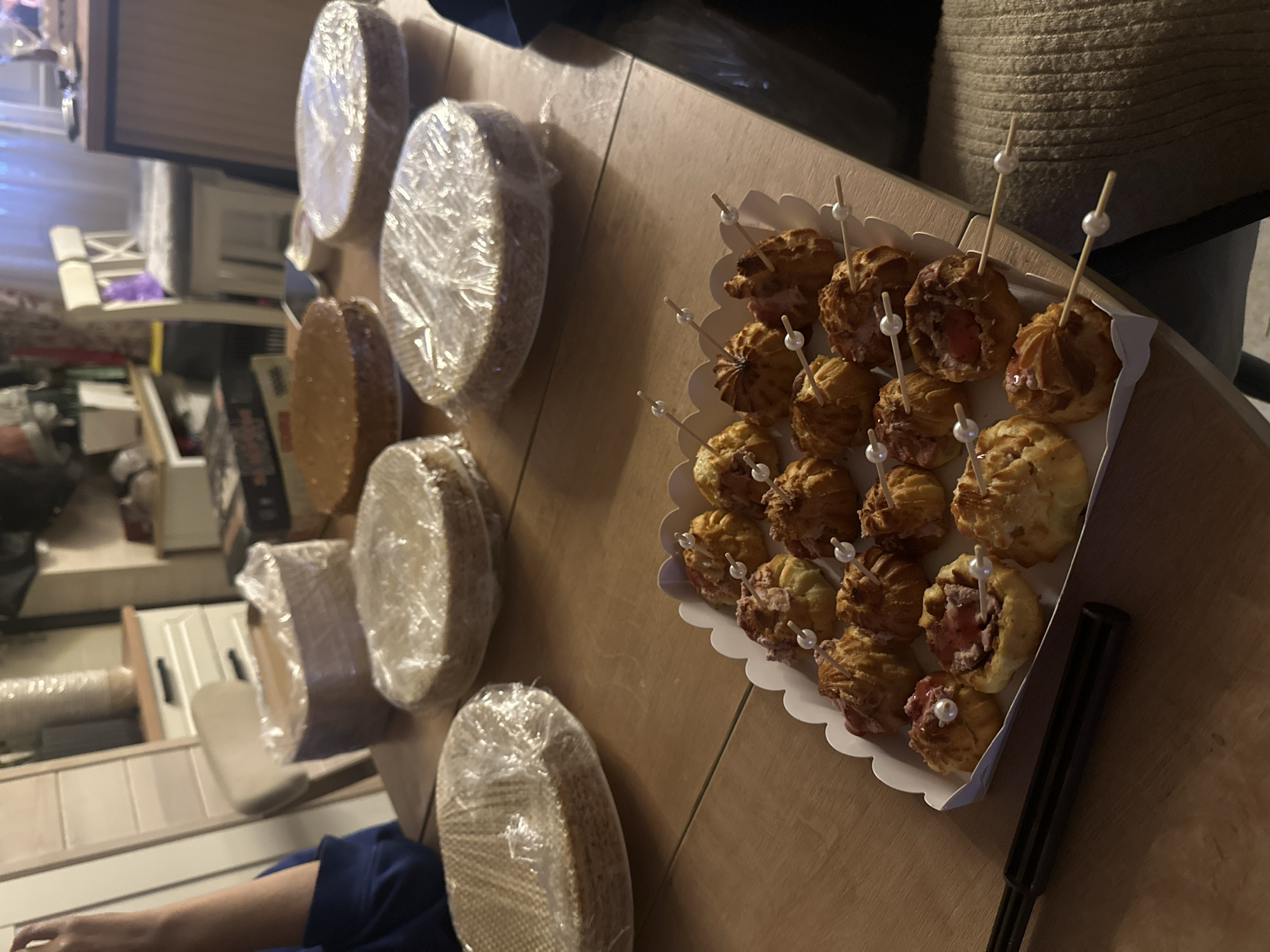 The image depicts a table laden with food items, likely in a domestic setting in Тумілавіцкі сельскі Савет, Belarus. In the foreground, a white scalloped tray contains an assortment of small, golden-brown pastries, some topped with red jam and decorative white pearls. These pastries are skewered with wooden toothpicks, also adorned with pearls. A black utensil rests to the left of the tray. In the background, several round cake bases are stacked and wrapped in clear plastic. One unfrosted cake base is visible without plastic. A person's arm, wearing a dark blue garment, is partially visible on the left side of the image. The table is made of wood, and the environment suggests an indoor scene, possibly a kitchen or dining area, with furniture and cabinets visible in the blurred background. The lighting is dim, creating a warm, ambient atmosphere.