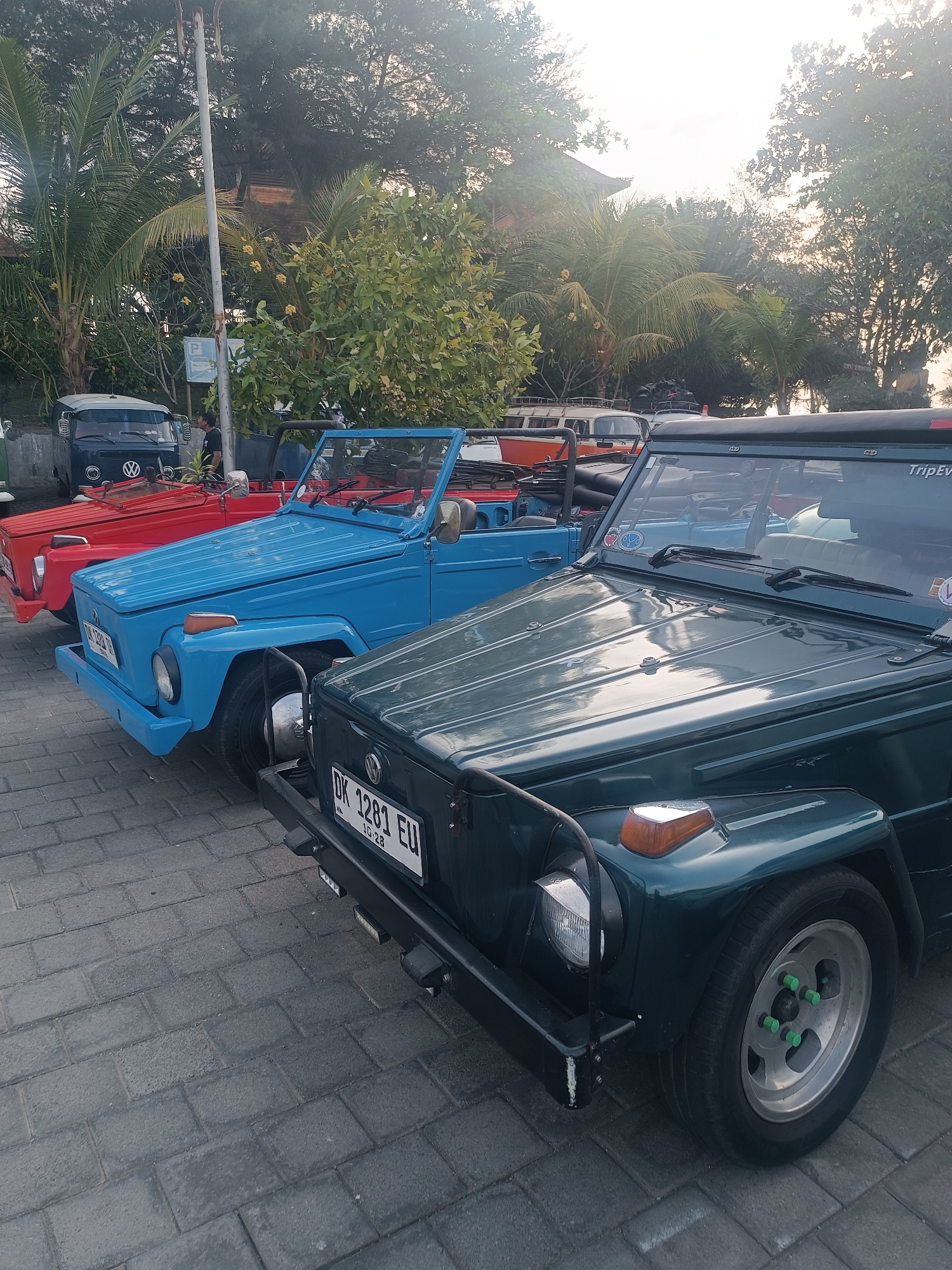 In Kediri, Indonesia, a row of vintage Volkswagen Thing vehicles is parked on a paved surface. The vehicles are arranged in parallel, with the closest being a dark green model. The dark green car has a black bumper, a visible license plate reading "DK 1281 EU 10-28", and a silver wheel with green lug nut covers. Behind it is a bright blue Volkswagen Thing with a black soft-top. The blue car has a chrome bumper and a license plate that is partially visible. Further back, a red Volkswagen Thing is parked, also with its soft-top down. In the background, a dark blue Volkswagen Type 2 van is visible. Palm trees and other lush green foliage surround the parking area, and a light pole stands near the side. The sky is bright and appears to be during the daytime, with some diffused sunlight.