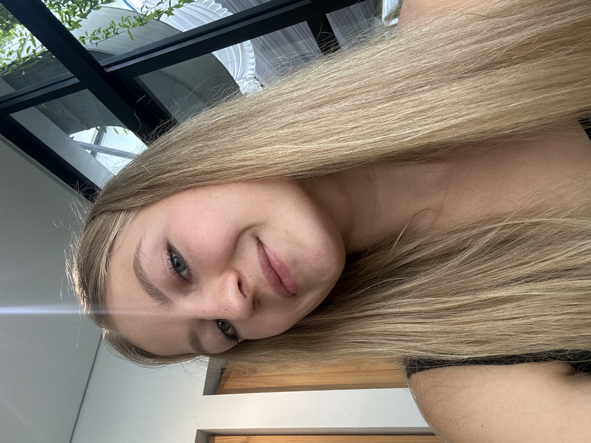 A selfie of a fair-skinned individual with long, blonde hair is angled to the left. The individual's face, featuring light eyebrows, gray eyes, a small nose, and pink lips, is partially obscured by their hair. In the background, a black-framed glass ceiling is visible, allowing for the observation of foliage. The individual appears to be indoors, with elements of wooden furnishings and a white wall discernable behind their shoulder. It is currently not possible to determine if this image was taken in Kuta Selatan, Indonesia.