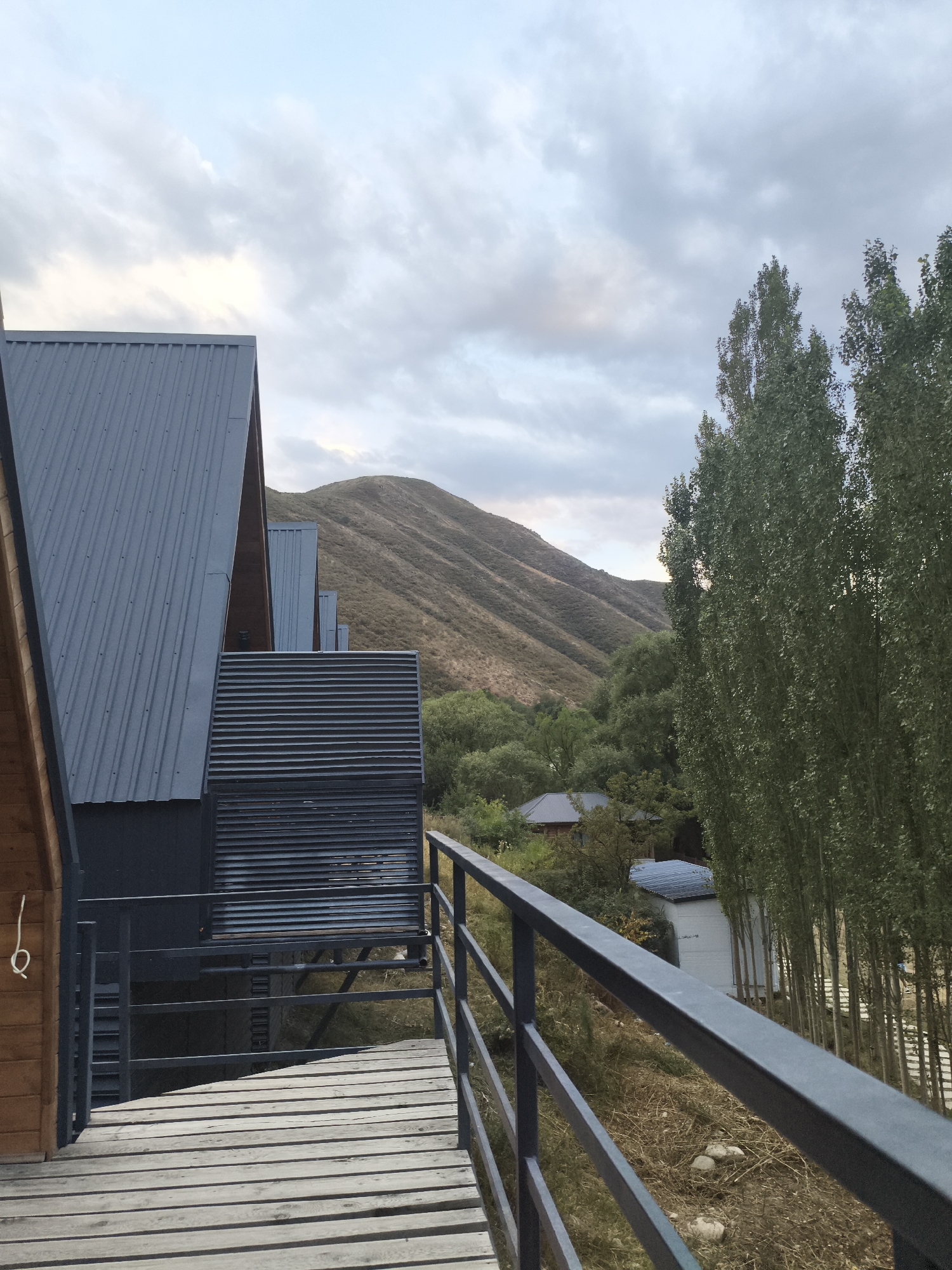 The image captures a view from a wooden boardwalk, looking towards a cluster of modern, angular buildings with dark metal roofs. The architecture is characterized by sharp lines and corrugated metal siding. In the background, a vast, undulating mountain landscape dominates the scene, its slopes covered in dry grass and sparse vegetation. A dense line of tall, slender trees runs parallel to the buildings on the right side of the frame. The sky is overcast with a mix of grey and white clouds, suggesting either early morning or late afternoon light. The overall impression is one of a modern retreat nestled within a natural, mountainous environment. The scene is located in Koy Tash, Kyrgyzstan.