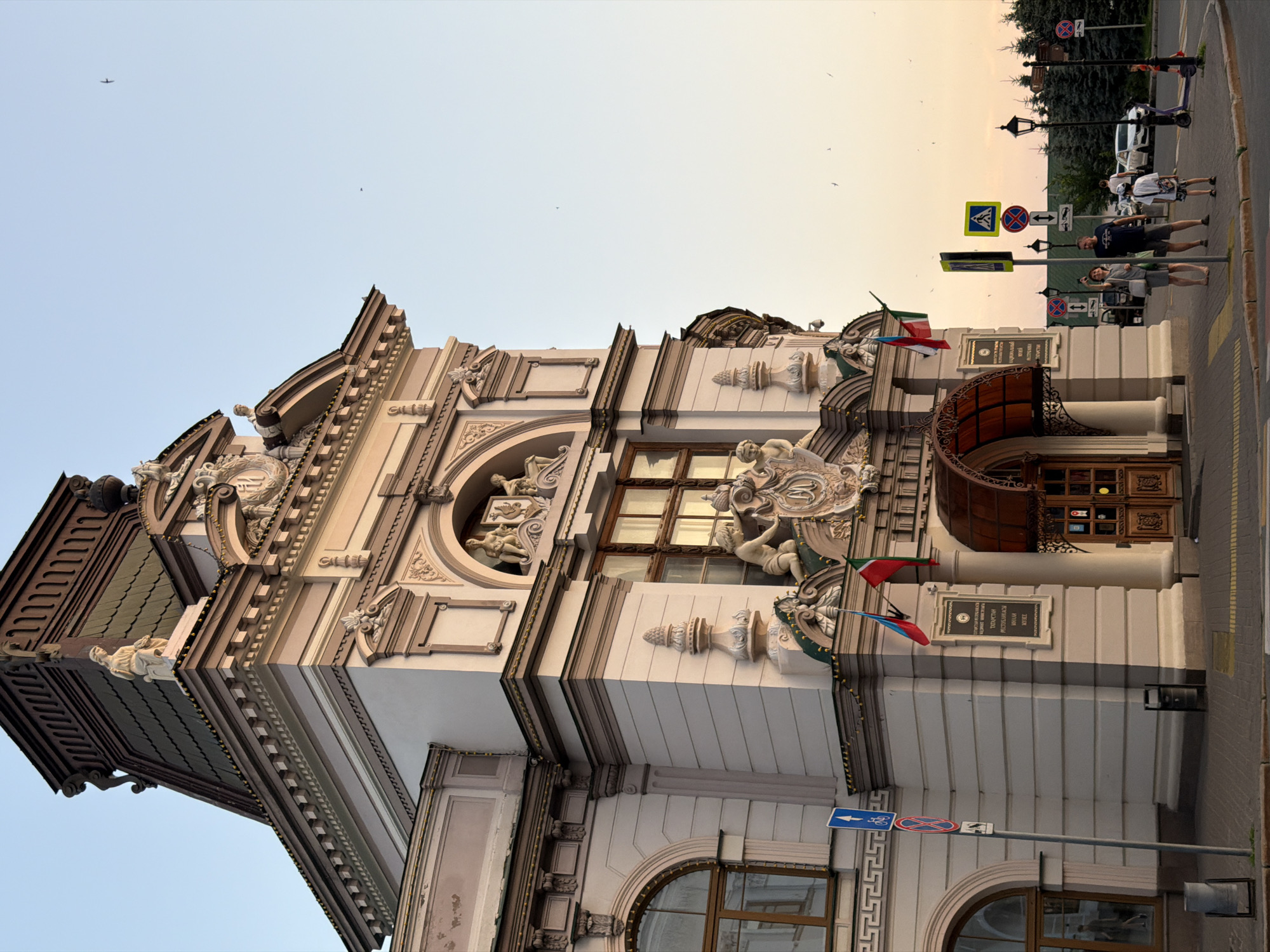 This is an exterior, low-angle shot of the National Museum of the Republic of Tatarstan in Kazan, Russia, bathed in the warm light of what appears to be late afternoon or early evening. The building's ornate facade features classical architectural elements, including statues of cherubs, detailed carvings, and a prominent arched entrance with wooden doors. Two flags, one red and one red, white, and green, are flown from the building. A man and a woman are standing on the sidewalk in front of the museum, the woman pointing upwards, possibly at the sky. Several other people and a few vehicles are visible in the background, along with street signs, lampposts, and trees. The sky is a clear, pale blue, with a few birds in flight.