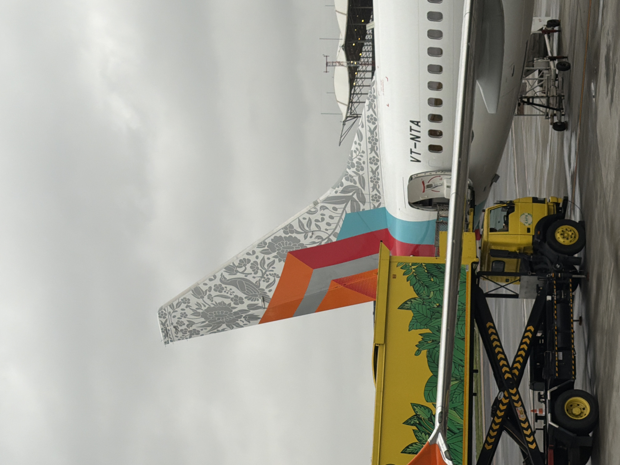 An airplane with a white fuselage and a decorative tail is parked on an airport tarmac. The tail features a grey floral pattern and orange, red, and turquoise geometric shapes. The text "VT-NTA" is visible on the fuselage. In the foreground, a yellow scissor lift truck is positioned next to a yellow cargo loader adorned with green leaf graphics. The truck has black and yellow chevron markings on its lifting mechanism. The scene is set against a cloudy sky, and in the background, airport structures with white roofs and a control tower are visible. The image appears to have been taken at Rajiv Gandhi International Airport in Hyderabad, India, given the typical aircraft registration prefix "VT" which is allocated to India and the presence of airport infrastructure. There are no people visible in the image.