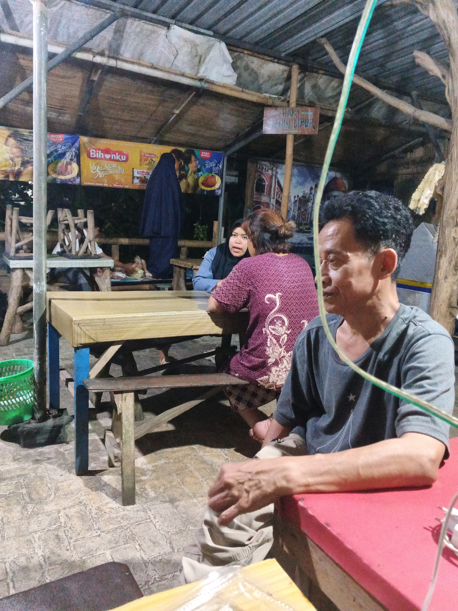 The image shows an outdoor dining area at night, possibly a warung or a street-side eatery in Ungaran, Indonesia. The scene is illuminated by artificial lights and features a rustic, makeshift roof supported by bamboo poles. Wooden tables and benches are present, with one table in the foreground covered in a light-colored wood grain pattern and supported by blue and brown legs. A green plastic basket sits on the ground near the table.
Several individuals are visible. In the foreground, a man in a dark grey t-shirt is seated and looking to the right. His right arm rests on a red surface in front of him. Behind him and to the left, a woman wearing a dark purple patterned shirt with white floral designs is seated at a table, looking towards the left. Next to her, a person in a dark blue headscarf is also seated. Further in the background, other figures can be seen sitting at tables, with some appearing to be obscured.
Banners with text and images are hung on the wall behind the seated individuals. One banner is yellow with red accents and features text in Indonesian, including "Bihunku" and "Rasa Ngamanku". Another banner, a darker color with lighter text, is also visible. The floor is paved with what appears to be textured tiles or concrete. The overall atmosphere suggests a casual dining environment.