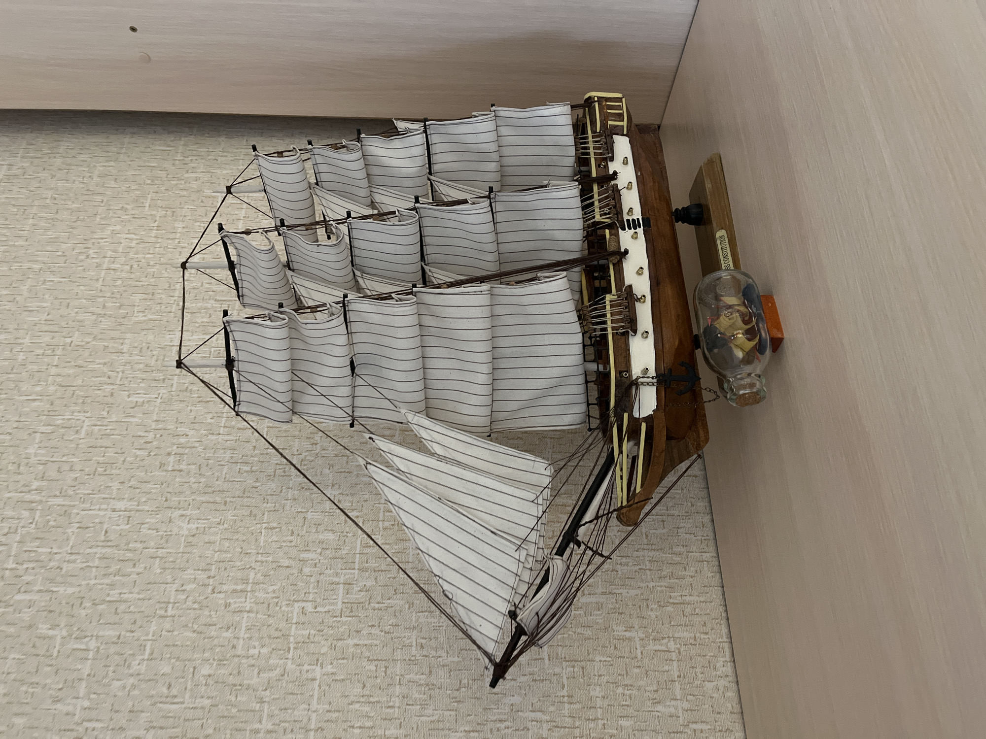 The photograph, taken in Chaikovsky, Russia, presents a detailed model of a sailing ship displayed indoors. The model is oriented vertically, appearing to sail upwards, likely resting on a shelf or mounted in a way that suggests this perspective.
The primary subject is a large, intricately crafted model of a multi-masted tall ship. Its sails are white fabric with fine, horizontal grey stripes, and they are furled or gathered, showcasing the extensive rigging made of thin, dark lines. The ship's hull and deck feature wooden elements, visible on the stern section with small details that appear to be miniature railings, cleats, and possibly cannons.
To the right of the ship, a small, clear glass bottle rests on a wooden block. The bottle contains what looks like sand and possibly a miniature item, resembling a "ship in a bottle" or a similar decorative maritime theme. On the side of the wooden block supporting this bottle, the text "USS CONSTITUTION" is clearly visible, suggesting the model ship might represent the famous American frigate or is displayed alongside memorabilia related to it.
The background consists of two distinct surfaces. On the left, there is a light beige or off-white wall covered with textured wallpaper, featuring a repeating, subtly raised brick-like pattern. On the right and extending across the top, there is a smooth, plain light-colored surface, possibly a ceiling or an adjacent wall. The lighting appears to be ambient indoor light, casting soft shadows. There is no activity taking place as it is a static display.