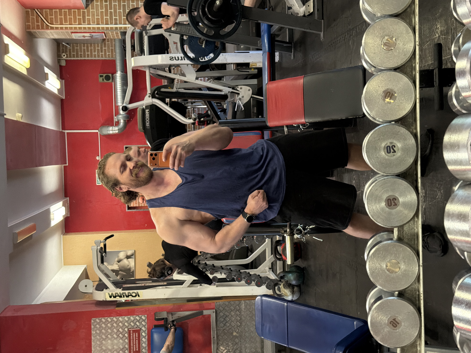 The image shows an interior gym setting. A man is taking a selfie, flexing his bicep while standing in front of a dumbbell rack. The dumbbells appear to be marked in kilograms. Gym equipment, including weight machines and benches, are visible in the background. Another person is using a machine in the background. The gym has red and white walls and standard gym lighting. It is impossible to determine if this is Saint Petersburg, Russia based solely on this image.