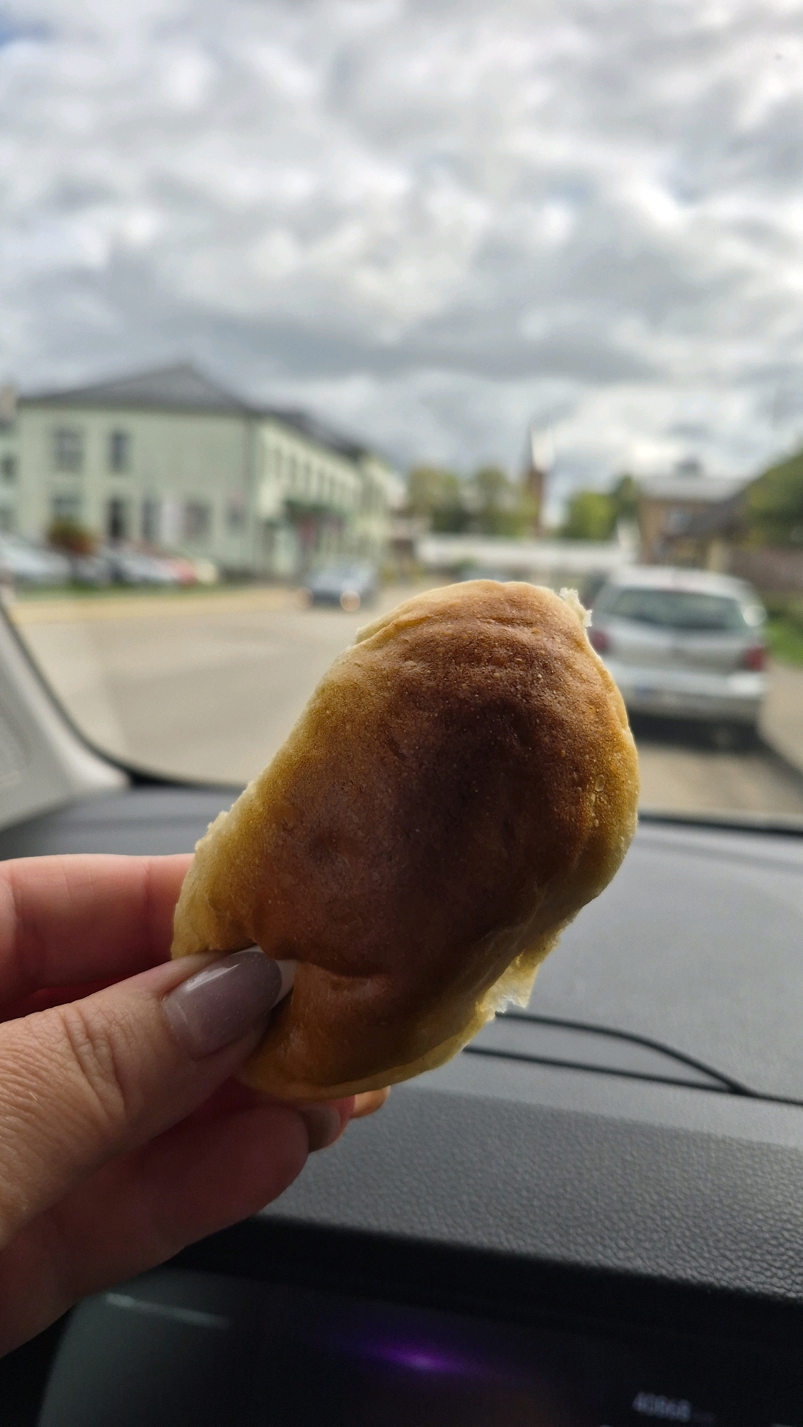 A hand with light-colored fingernail polish is holding a small, golden-brown baked good, possibly a pastry or roll, in front of a car's dashboard. The background, visible through the car's windshield, shows a slightly overcast sky with clouds. In the middle distance, there are buildings and parked cars, suggesting an urban or town setting. The architecture of the buildings appears to be of a European style, with pitched roofs and multiple stories, consistent with the town of Preiļi, Latvia. A light-colored car is parked on the right side of the street. The overall impression is a casual snapshot taken from inside a vehicle, focusing on the food item.