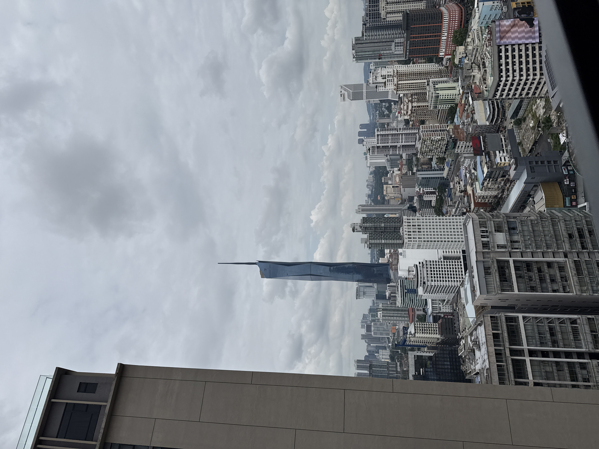 The image shows an aerial view of Kuala Lumpur, Malaysia, under a cloudy sky. Dominating the skyline is the Merdeka 118 skyscraper, recognizable by its pointed, triangular spire. Below are dense clusters of buildings of varying heights and architectural styles, typical of an urban environment. A low building with a horizontal design is visible in the lower left corner of the frame. The lighting is muted, suggesting an overcast day.