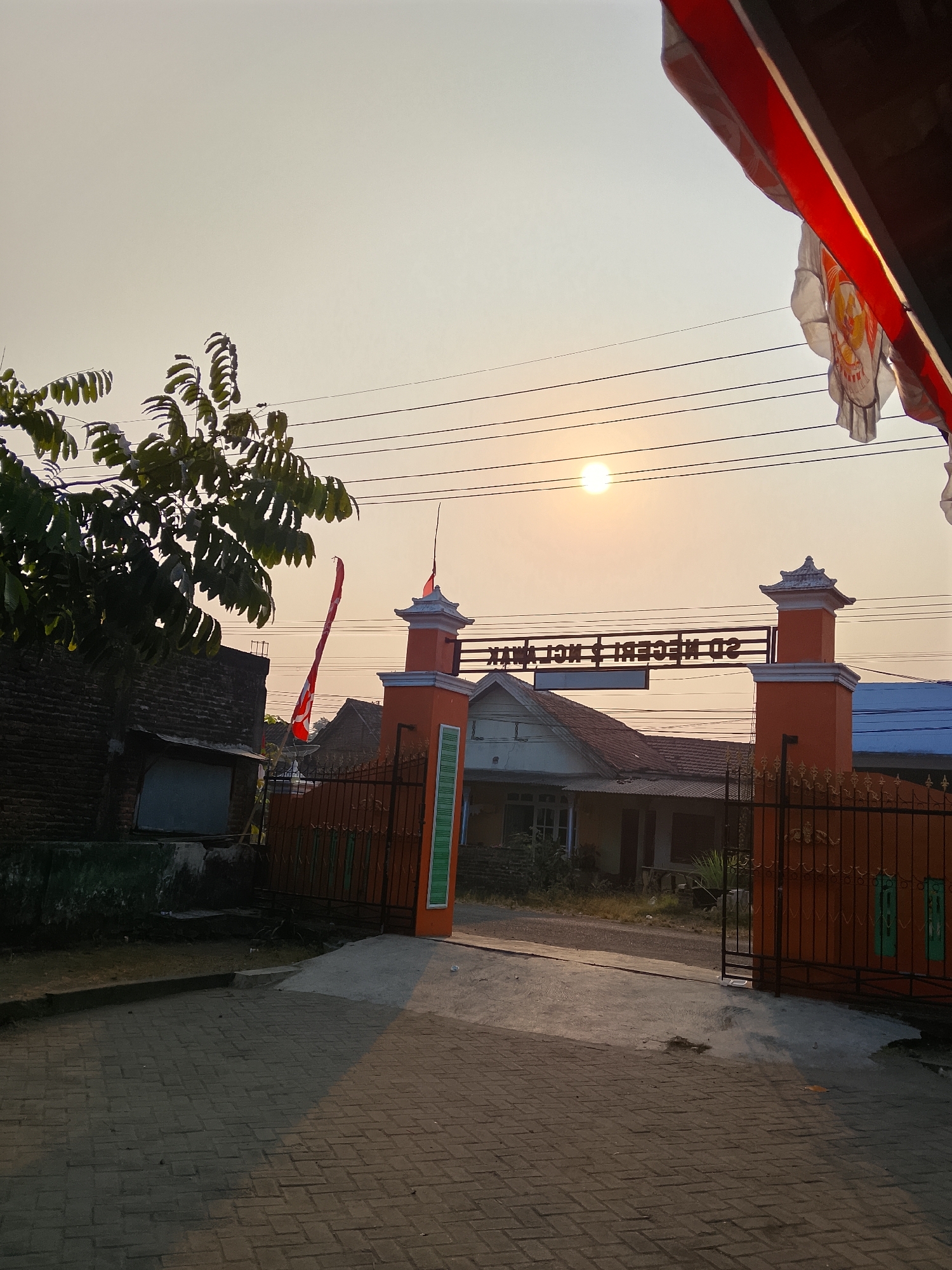The image shows the entrance to an Indonesian school in Kepuh, Indonesia, at sunset. The sun is low in the sky, casting a warm glow over the scene. The entrance is marked by an orange archway with a sign that reads "SD NEGERI 02". The archway is flanked by two pillars, and a metal gate is visible in front of the entrance. To the left of the entrance is a brick building, and to the right, a white building with a blue roof. A tree with green leaves is on the left side of the image. Overhead, power lines crisscross the sky, and to the right, a red and white Indonesian flag is hanging from a building. The ground is paved with interlocking bricks. The scene is peaceful and serene, capturing the quiet beauty of a rural Indonesian village at dusk.