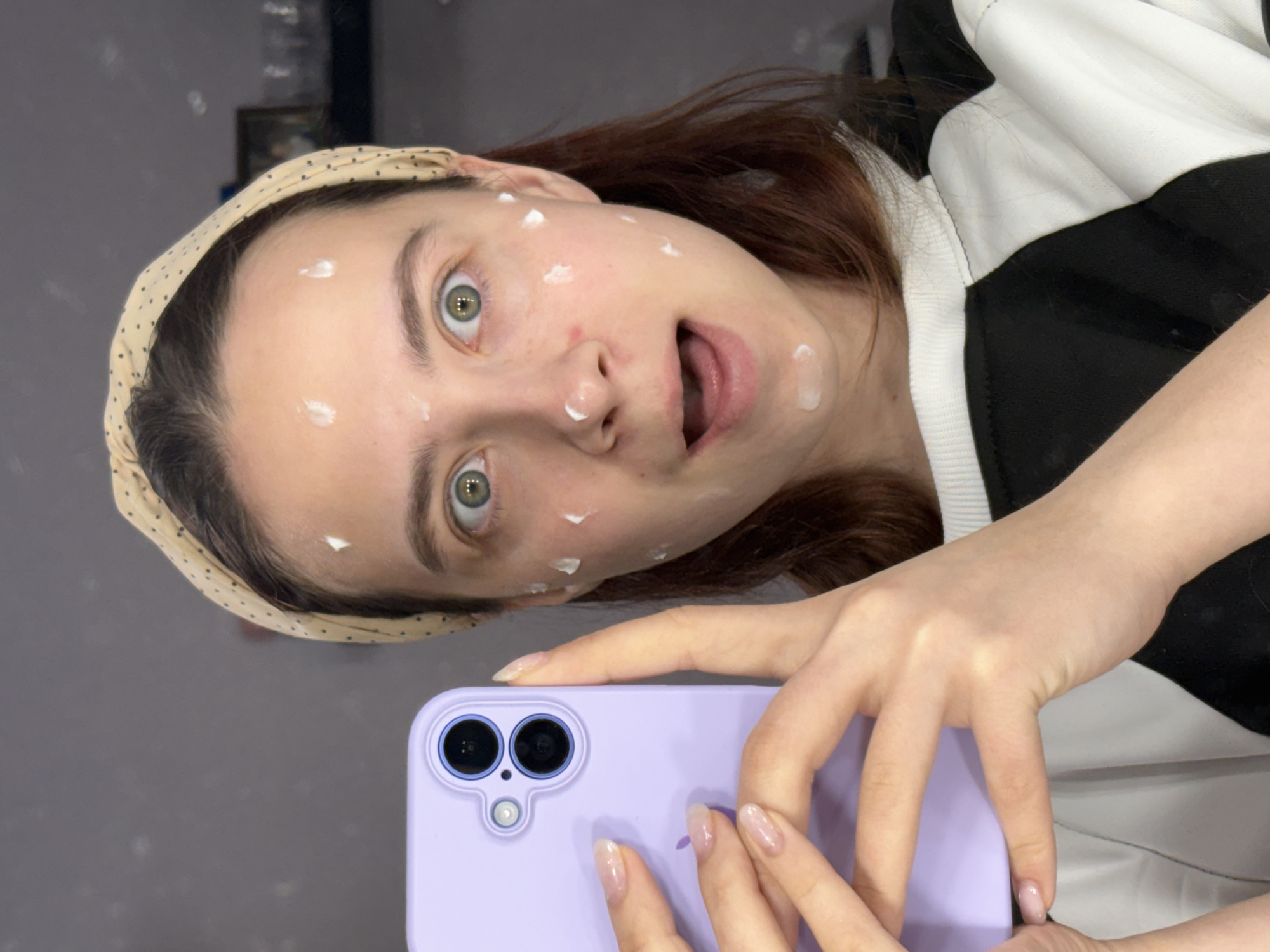 A woman is taking a selfie with a purple smartphone. She is wearing a dotted headband and a black and white shirt. There are white cream spots on her face. The background appears to be a grey wall. The location of Прикамский, Russia, is not discernible from the image.
