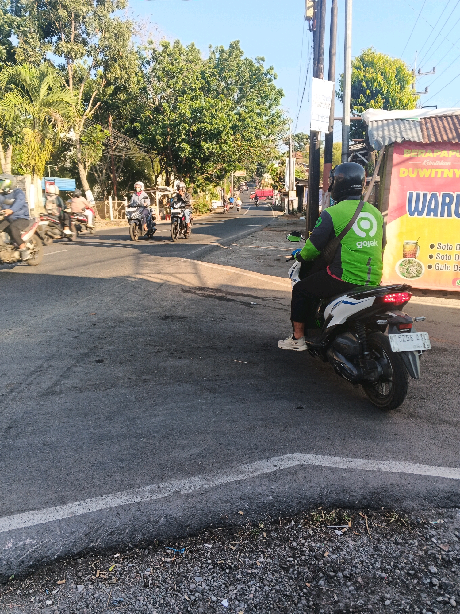 The image depicts a street scene in Semarang, Indonesia, on a sunny day. A person wearing a green Gojek jacket and helmet is riding a white motorcycle. The motorcycle has a license plate reading "H 5256 ASC". In the background, other motorcycles and people are visible on the road, with trees lining the street. A building with a red and yellow sign advertising "WARUNG SOTO DOWITNYA" is also present on the right side of the frame. The road surface is asphalt with a white line marking a lane. The area adjacent to the road is unpaved and contains gravel and debris.