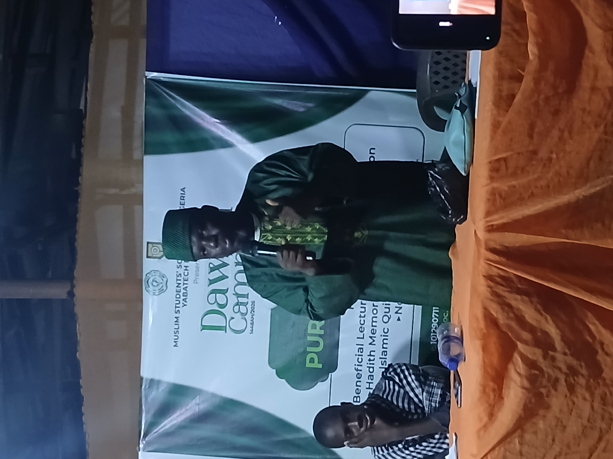This image depicts an indoor event in Lagos, Nigeria. A man in a green traditional outfit and cap is speaking into a microphone. He is standing behind a table covered with an orange cloth. To his left, a young man in a checkered shirt is seated, resting his chin on his hand. A banner in the background features text that includes "MUSLIM STUDENTS' SO," "YABATECH," "Daw," "CAMPA," and "NIGERIA." Other items visible on the table include a plastic water bottle, a smartphone, and a black bag. The environment appears to be dimly lit.