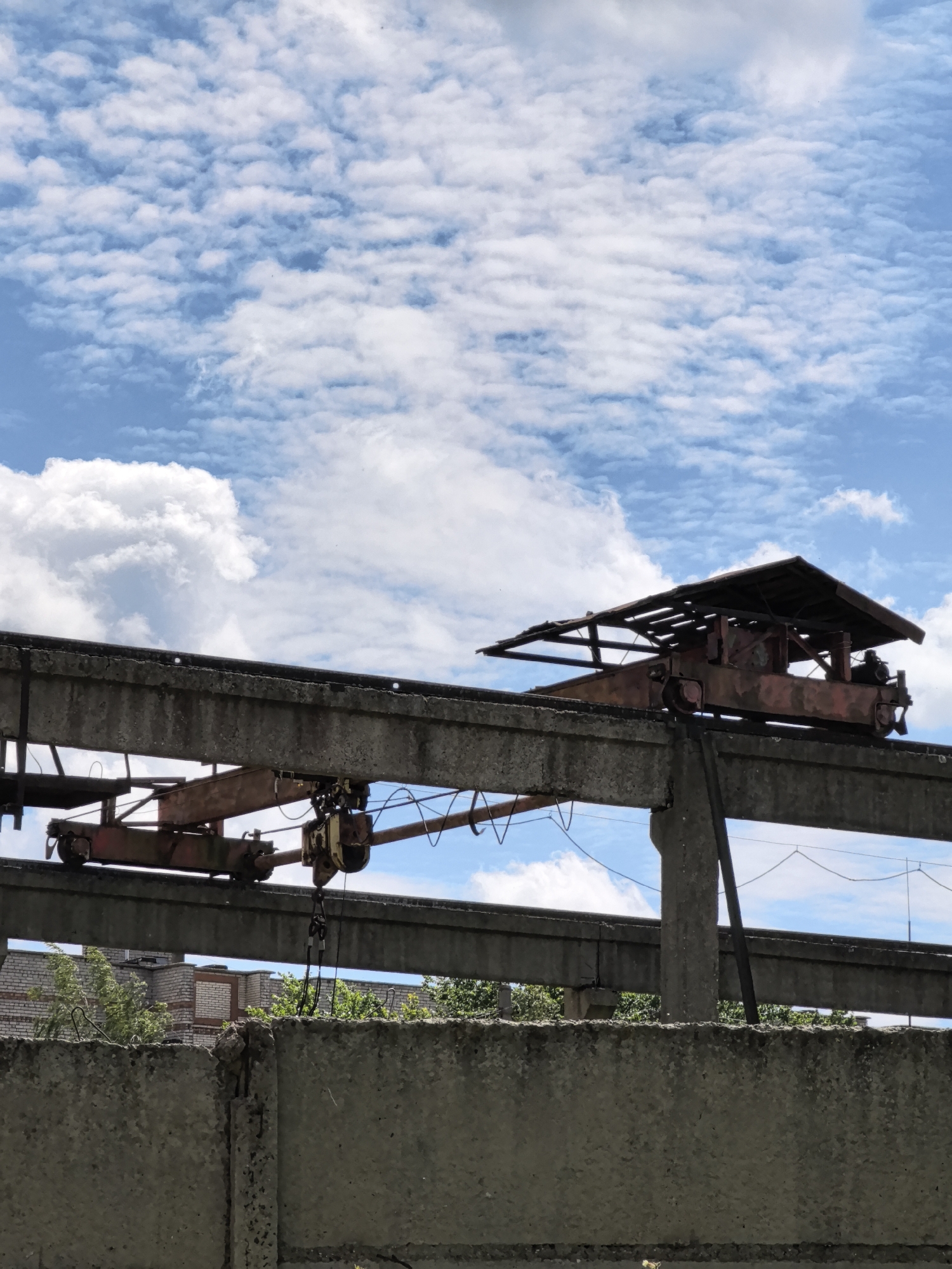 An outdoor, medium shot captures a bright, cloudy day with a clear blue sky. In the foreground, a weathered concrete wall runs horizontally. Above the wall, two large, parallel concrete beams are supported by concrete pillars. These beams form an overhead structure, possibly part of an industrial site. A rusted overhead crane, with a partially damaged roof, is visible on the upper beam, positioned to the right of the frame. Another, smaller crane or trolley mechanism is attached to the lower beam, to the left. Vegetation, including trees and shrubs, grows beneath the beams, and a brick building is partially visible in the background. The scene conveys a sense of abandonment or disuse.