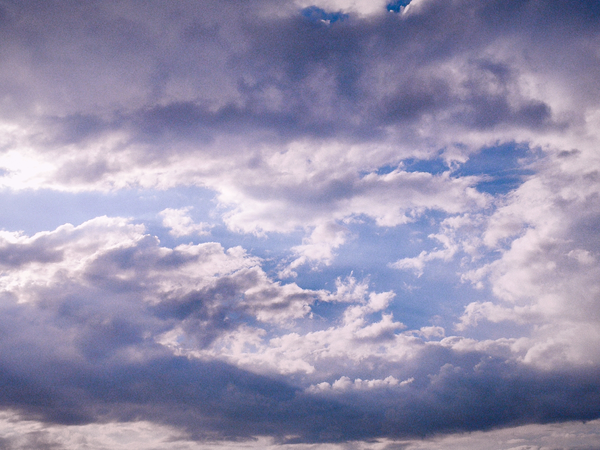 A low-angle, wide shot captures a dramatic sky filled with a mix of dark, purple-tinged clouds and bright, white cumulus clouds. Patches of blue sky are visible between the cloud formations. The lighting suggests it could be late afternoon or early morning, with sunlight filtering through the clouds, creating highlights and shadows. There are no people, structures, or other discernible objects in the image, making the location impossible to determine.