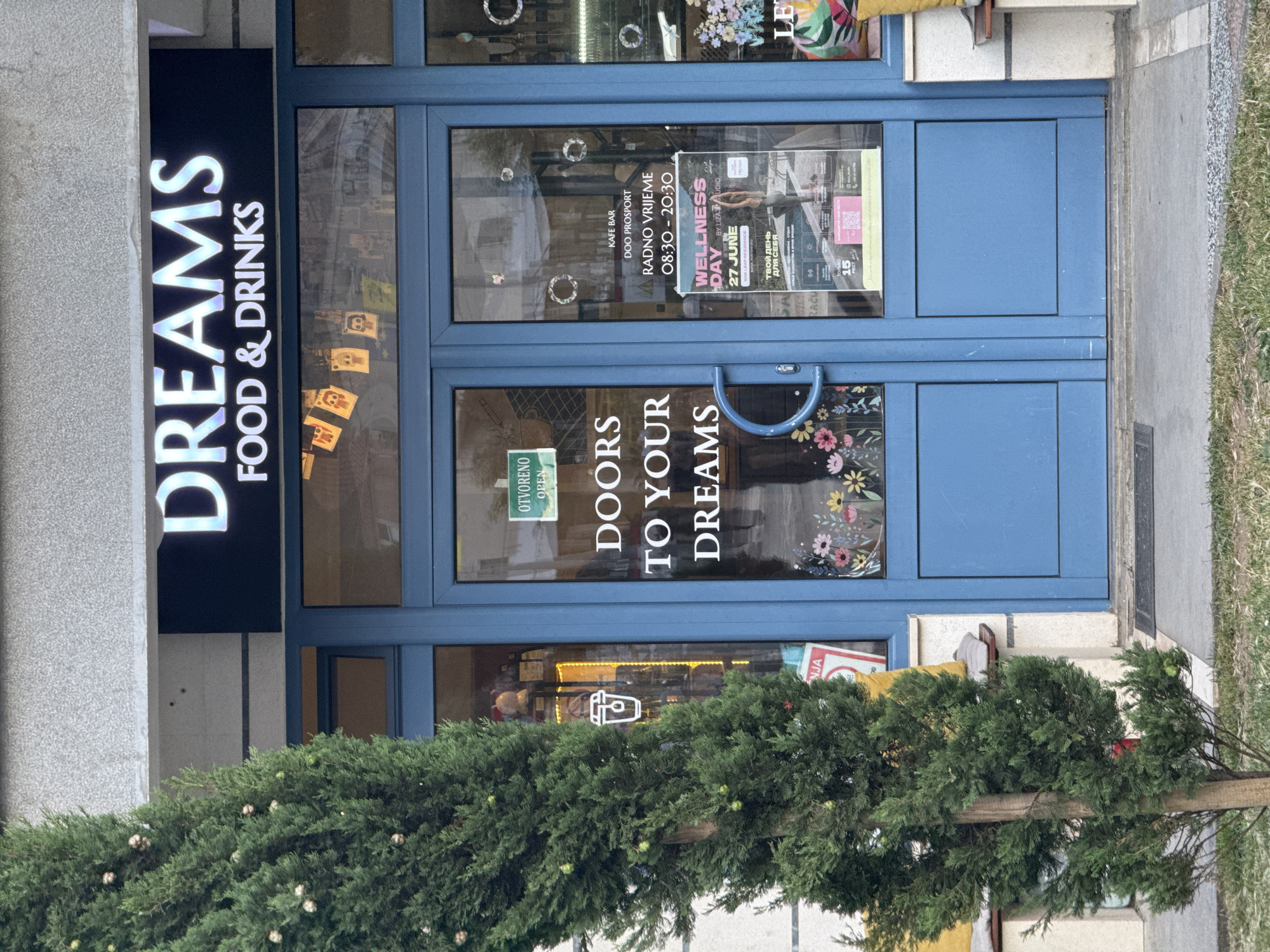 The image shows the exterior of a shop with a blue door and a large window displaying text that reads "DREAMS FOOD & DRINKS". A tree stands in front of the shop. The door has a sign that says "OTVORENO OPEN" in green. Inside the window, there is a poster advertising a "WELLNESS DAY" event on June 27th. The shop appears to be located in Budva, Montenegro, as indicated by a sign on the door with the text "NJAZA" which is likely a reference to the popular beach in Budva. The overall scene is well-lit, suggesting it was taken during the daytime.