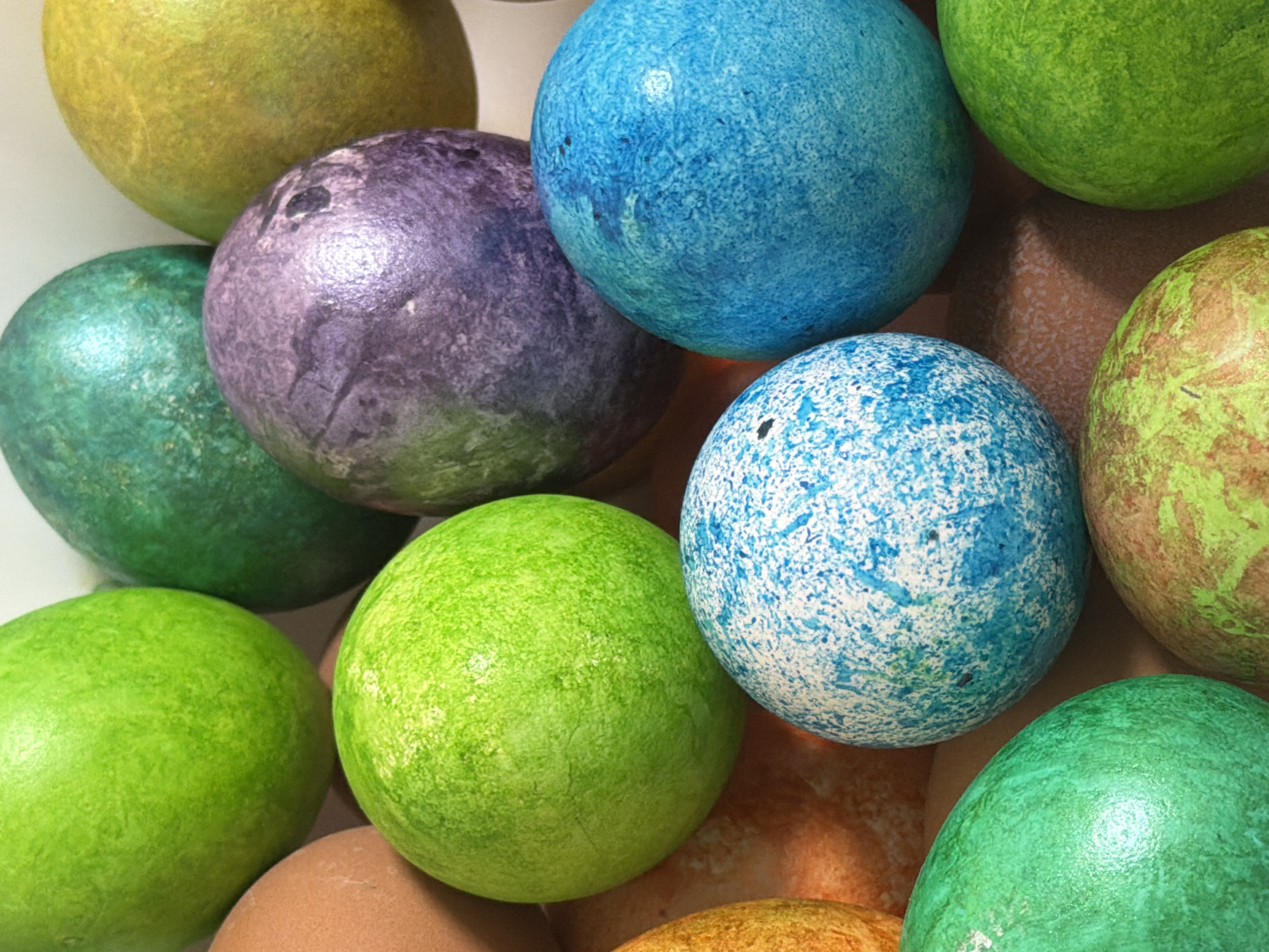 The image shows a close-up, high-angle view of approximately a dozen decorated Easter eggs. The eggs are in a variety of colors including shades of green, blue, yellow, and purple. The eggs are clustered together, partially overlapping each other. The surfaces of the eggs feature various patterns and textures. The eggs are resting on a surface that is out of focus. There is no indication of the location in the city of Chaikovsky, Russia.