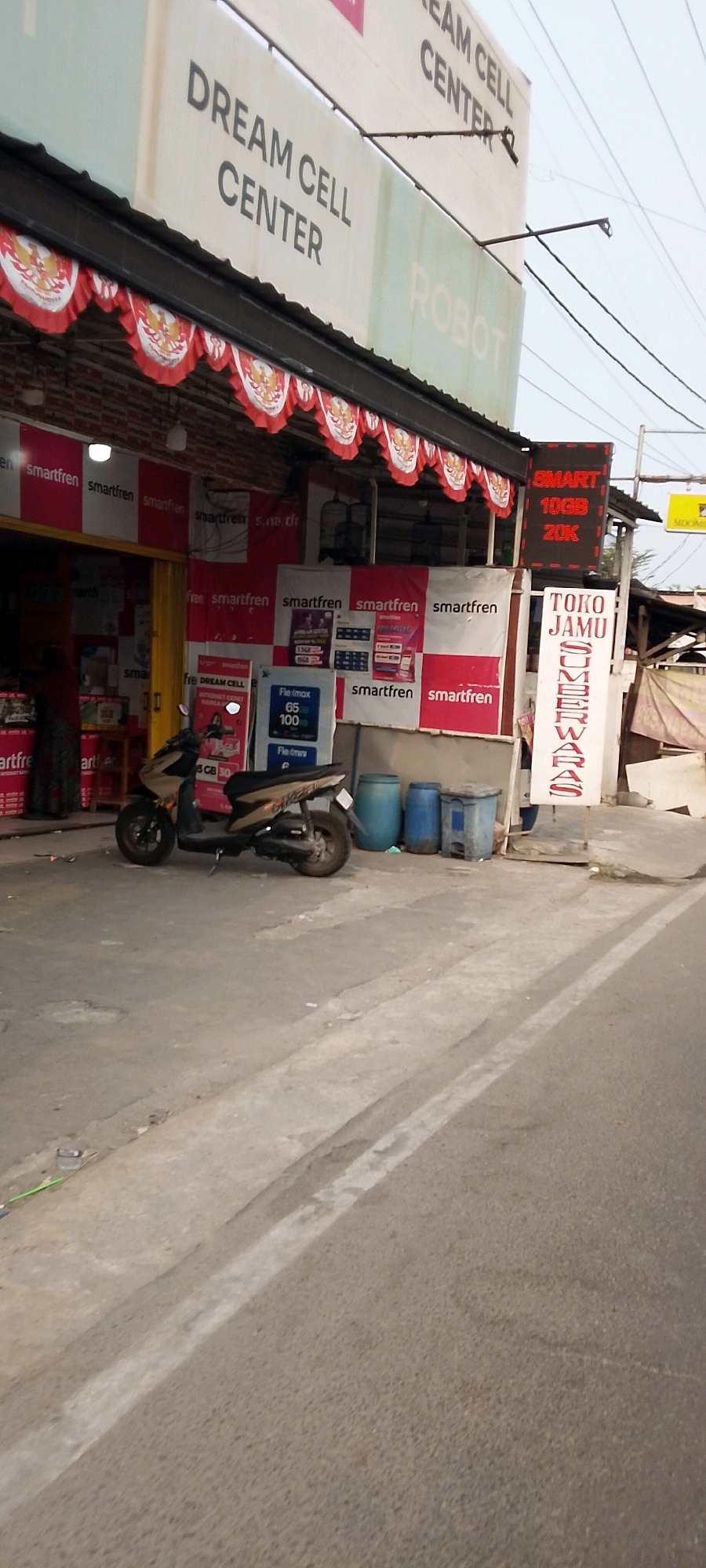 The image depicts a street-level view of a retail establishment in Sriamur, Indonesia. The prominent business is "DREAM CELL CENTER," as indicated by signage above the storefront. To the right, a red digital sign displays "SMART 10GB 20K," likely advertising a mobile data plan. Adjacent to this, a vertical sign reads "TOKO JAMU SUMBER WARAS," suggesting a traditional herbal medicine shop.
The storefront features red and white banners, some with the "smartfren" logo, indicating it's a telecommunications retailer. Several large blue plastic barrels are visible near the entrance, along with a grey trash bin. A beige and black scooter is parked on the asphalt in front of the shop. The ground is paved, with white painted lines indicating a road or pathway. Power lines are visible overhead. The overall scene suggests a commercial area with multiple small businesses. No people are clearly visible in the frame, but the presence of a parked scooter implies potential activity within the shops.