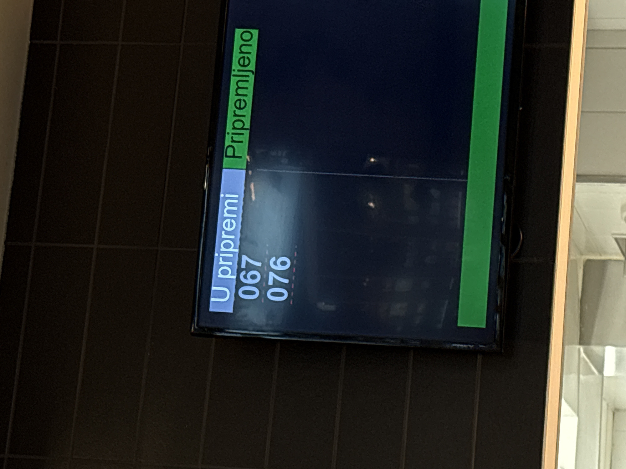 The image shows a digital display mounted on a dark, tiled wall. The screen indicates an order status in Serbian: "U pripremi" (in preparation) with order numbers "067" and "076," and "Pripremljeno" (prepared). The location is likely Kragujevac, Serbia based on the language of the display.