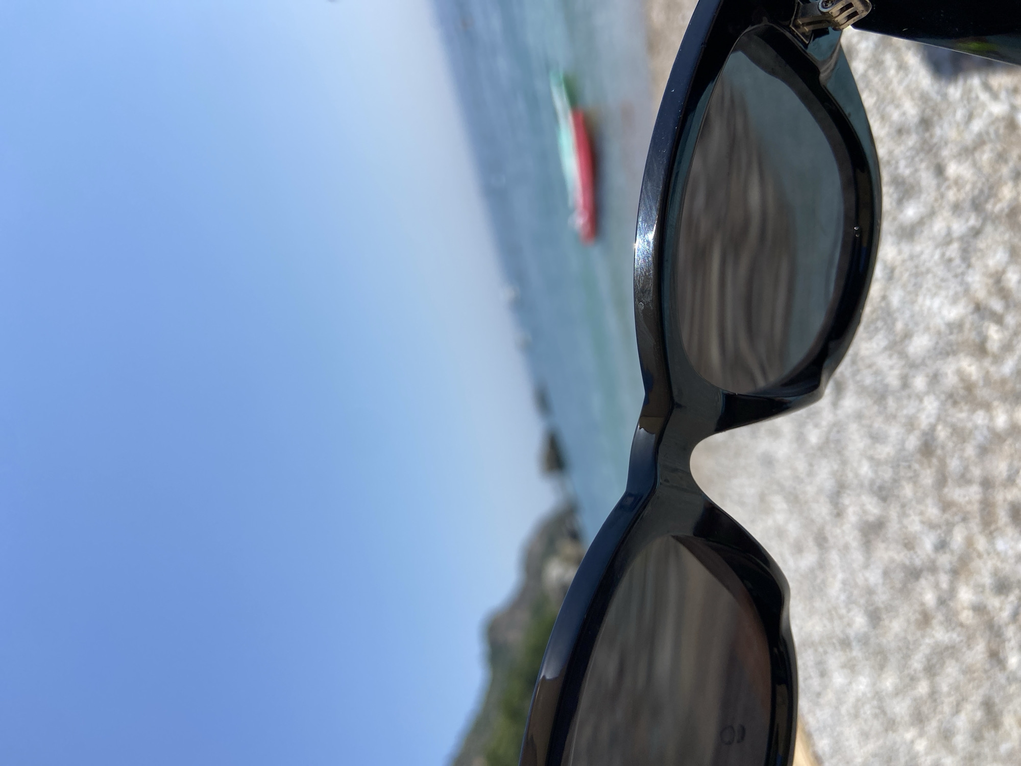 The image is a close-up, first-person perspective shot taken through a pair of black sunglasses. The sunglasses are the primary focus, with their frames and lenses filling the foreground. Beyond the sunglasses, a beach scene unfolds under a clear blue sky.
The beach appears to be covered in small, light-colored pebbles or gravel. In the middle ground, the turquoise water of the sea stretches out, meeting the horizon. Several small boats or buoys are visible on the water. One prominent inflatable object, colored red and green, floats on the surface of the sea.
In the background, a hilly, mountainous landscape rises from the coastline, partially obscured by a slight haze. The overall impression is a sunny day at a coastal location. While the specific location of Prijevor, Montenegro, is provided as context, the image itself does not contain any explicit markers or recognizable landmarks to confirm this specific geographical placement.