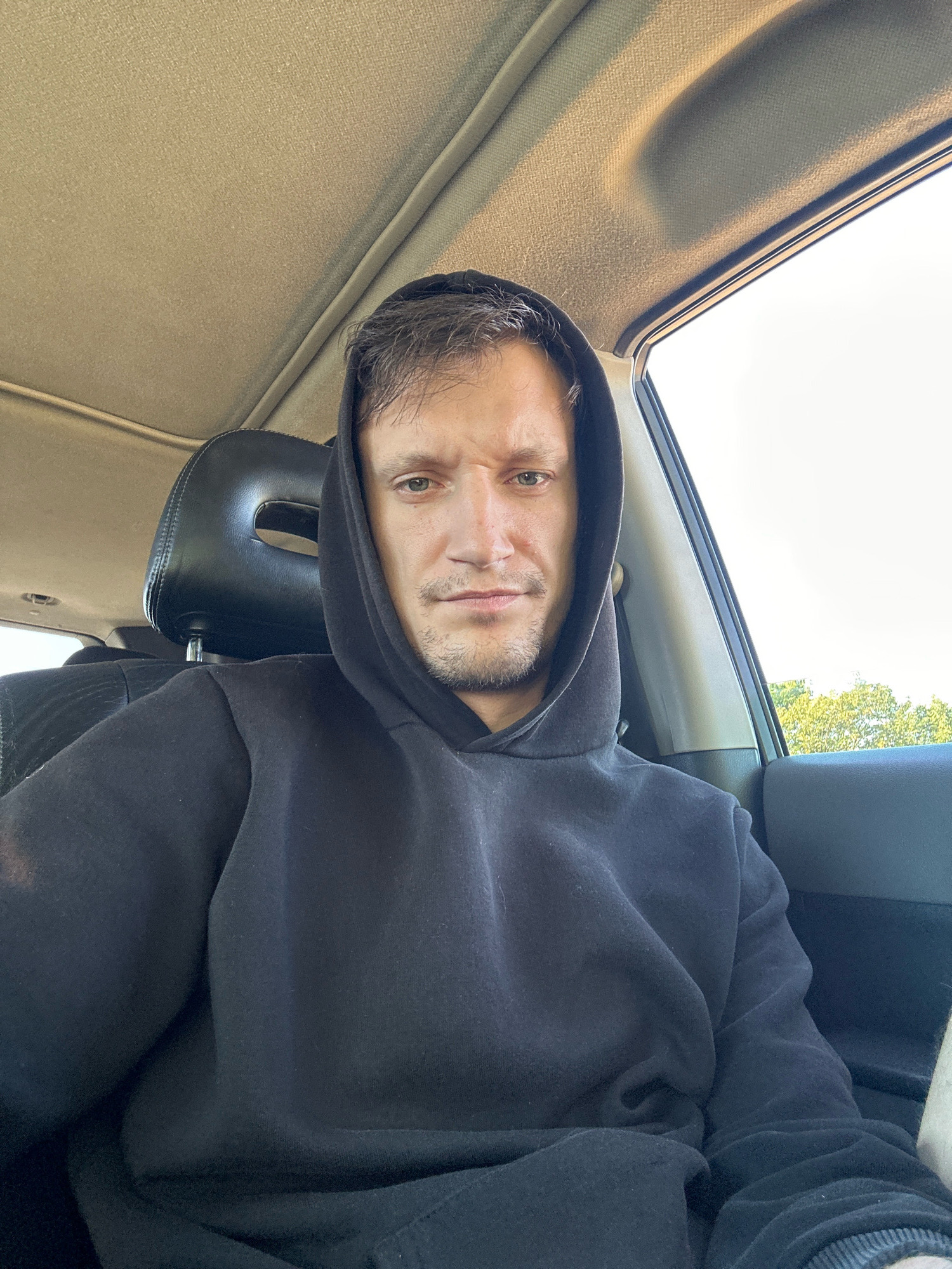 A medium shot captures a man from the shoulders up, sitting inside a vehicle. He is wearing a black hooded sweatshirt, with the hood pulled up and partially obscuring his head. His face is visible, turned towards the camera, with a neutral expression. The interior of the car is visible behind him, including a black headrest and the beige headliner of the ceiling. A window on the right side of the frame shows a blurred view of green trees and a bright, overcast sky. The lighting suggests it is daytime. The man's location is in the city of Makiv, Ukraine.