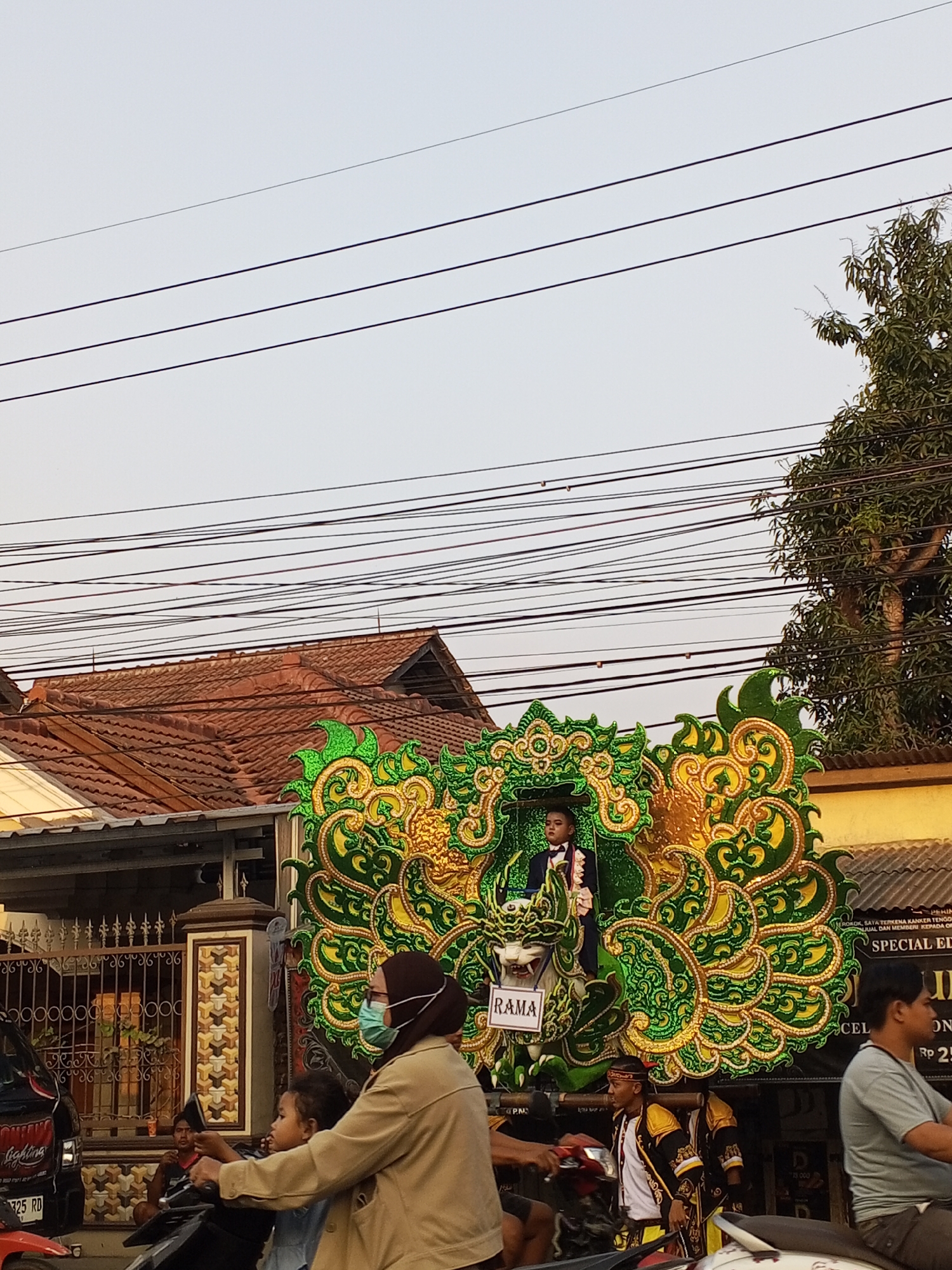 In Gebang, Indonesia, a parade float adorned with vibrant green and gold dragon-like motifs is depicted. A young boy, dressed in a formal suit with a bow tie, stands atop the float, appearing as if he is part of the performance. Below him, a sign reads "RAMA". Several individuals in traditional attire are also visible, some carrying the float, while others are on motorcycles, observing the procession. A woman wearing a face mask and a light brown jacket is seen on a motorcycle, accompanied by a child. In the background, residential buildings with tiled roofs and a dense network of power lines suggest a typical urban or semi-urban environment. The sky is overcast, and a large tree with dense foliage is visible on the right side of the image. The overall scene captures a cultural event or celebration taking place in the city.