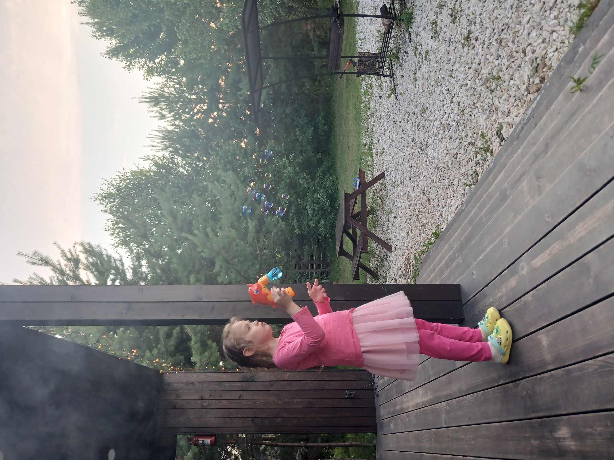 A young girl, wearing a pink long-sleeved shirt, pink leggings, and a pink tutu, stands on a dark wooden deck outdoors. She is holding a toy that appears to be a bubble gun in her right hand. Her left hand is raised. She is looking to her left. Yellow clogs with flower designs are on her feet. In the background, numerous colorful bubbles are floating in the air. Beyond the bubbles, there is lush green foliage and trees. To the right of the girl, a gravel area is visible, with a dark metal structure that looks like a grill gazebo. The sky above is overcast and appears to be late afternoon or early evening. The setting is likely a backyard or recreational area.