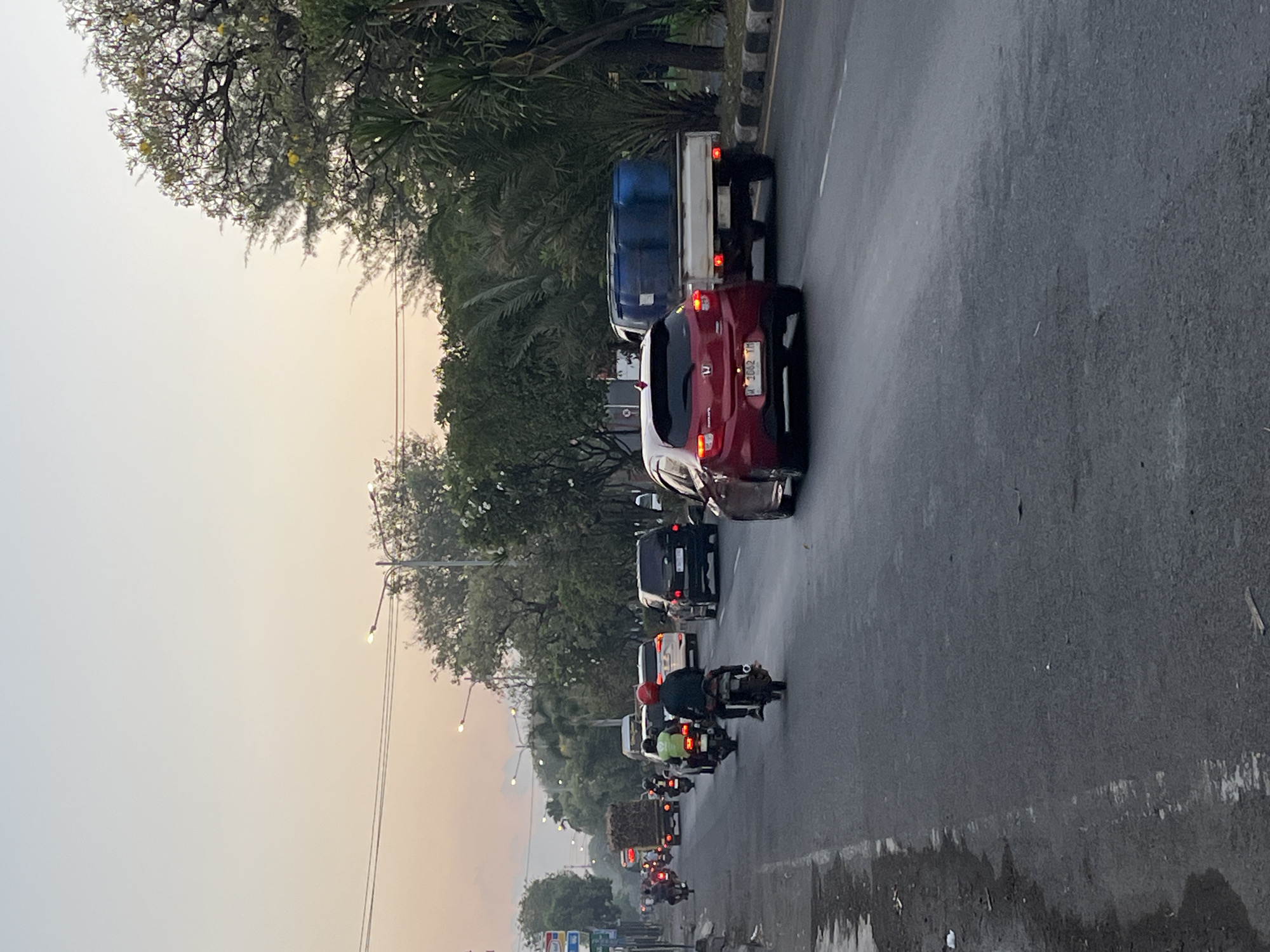 The image depicts a street scene in Ngaban, Indonesia, during what appears to be late afternoon or early evening. The sky is a soft gradient of pale blue and yellow, indicating a sunset or sunrise. Traffic is moderate, with a mix of cars and motorcycles.
On the right side of the road, a red hatchback car is in the foreground, followed by a white pickup truck carrying several large blue barrels. Further down the road, a black SUV and a white car are visible. On the left side of the road, several motorcycles are in motion. One motorcycle in the center has a rider wearing a red helmet. Another motorcycle further back has a rider in a green shirt. In the distance, a truck loaded with what appears to be hay or straw is moving away from the camera.
The road is paved and appears somewhat wet in places, with some debris scattered along the left shoulder. Trees line the right side of the road, with some streetlights visible along the median. The overall atmosphere is that of a busy, but not overly congested, urban thoroughfare during a transitional time of day.