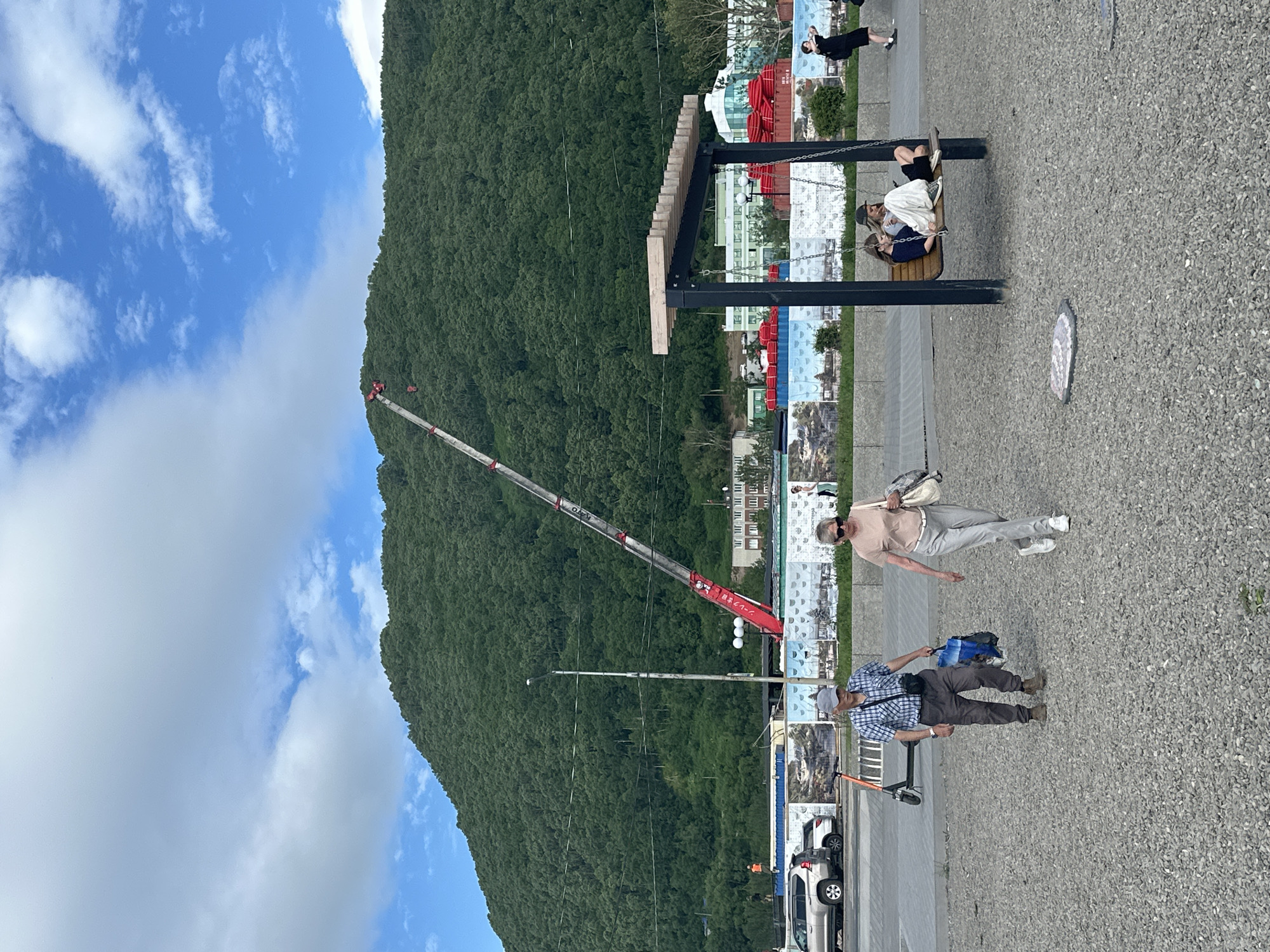This image depicts a daytime scene in Petropavlovsk-Kamchatsky, Russia, with a large, tree-covered hill dominating the background. A long, red and white crane extends diagonally from the lower left towards the top of the hill. In the foreground, a gravel-covered area features a black and wooden swing set where two young individuals are seated. To the left of the swing set, an older man wearing a plaid shirt and cap walks with a shopping bag in his hand, accompanied by an older woman in a light-colored shirt and pants. Behind them, a grey SUV and an orange electric scooter are parked. A series of posters or murals are visible on a low wall between the paved area and the background, featuring various scenes. Buildings and shipping containers are visible at the base of the hill. The sky is blue with scattered white clouds.