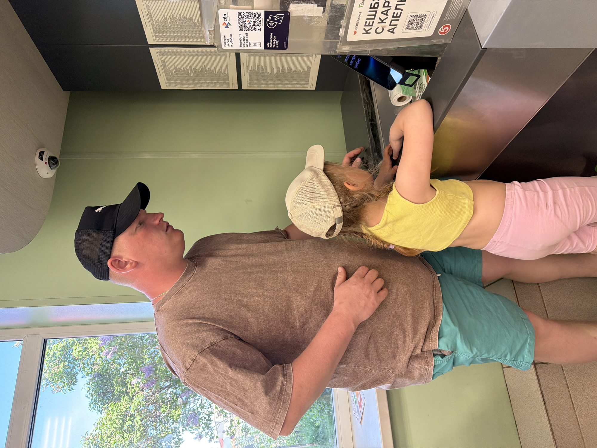 The image depicts a man and a child at an automated kiosk. The man wears a dark baseball cap, a brown t-shirt, and teal shorts. The child, wearing a light yellow top, pink leggings, and a tan baseball cap, reaches towards the kiosk screen.
The kiosk features printed information sheets and signage in Russian, including payment options like QR code and NFC. One sign mentions "Апельсин" (Orange) and "КЕШБ С КАР АПЕЛЬ" (Cashback with Orange Card), suggesting a loyalty or payment system. A surveillance camera is visible on the wall.
Through a nearby window, green foliage and trees in blossom are visible, suggesting an outdoor setting during a mild season. The location is likely a public space or business in the city of Chaikovsky, Russia, as inferred from the Russian signage on the kiosk.