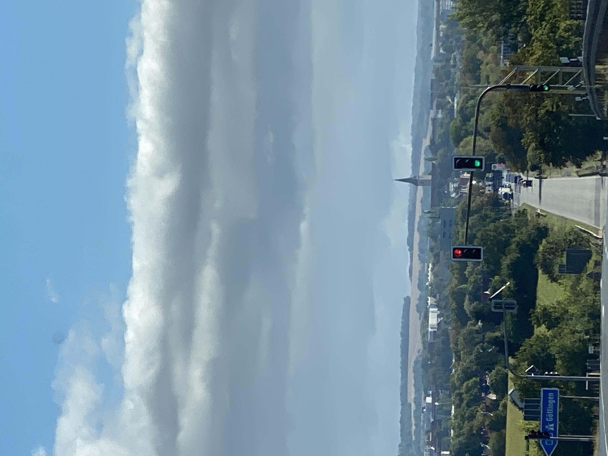 The image captures a view from an elevated position looking down a road lined with trees. In the foreground, a traffic light is red on the right and green on the left. To the left, a blue road sign with white lettering indicates "Göttingen". Further down the road, vehicles are visible, and another traffic light shows green. In the distance, a city skyline is present with a prominent church spire. The sky is partly cloudy with large, white clouds against a blue background. The overall scene suggests an outdoor setting in a German city, possibly Werther, Germany, given the road sign.