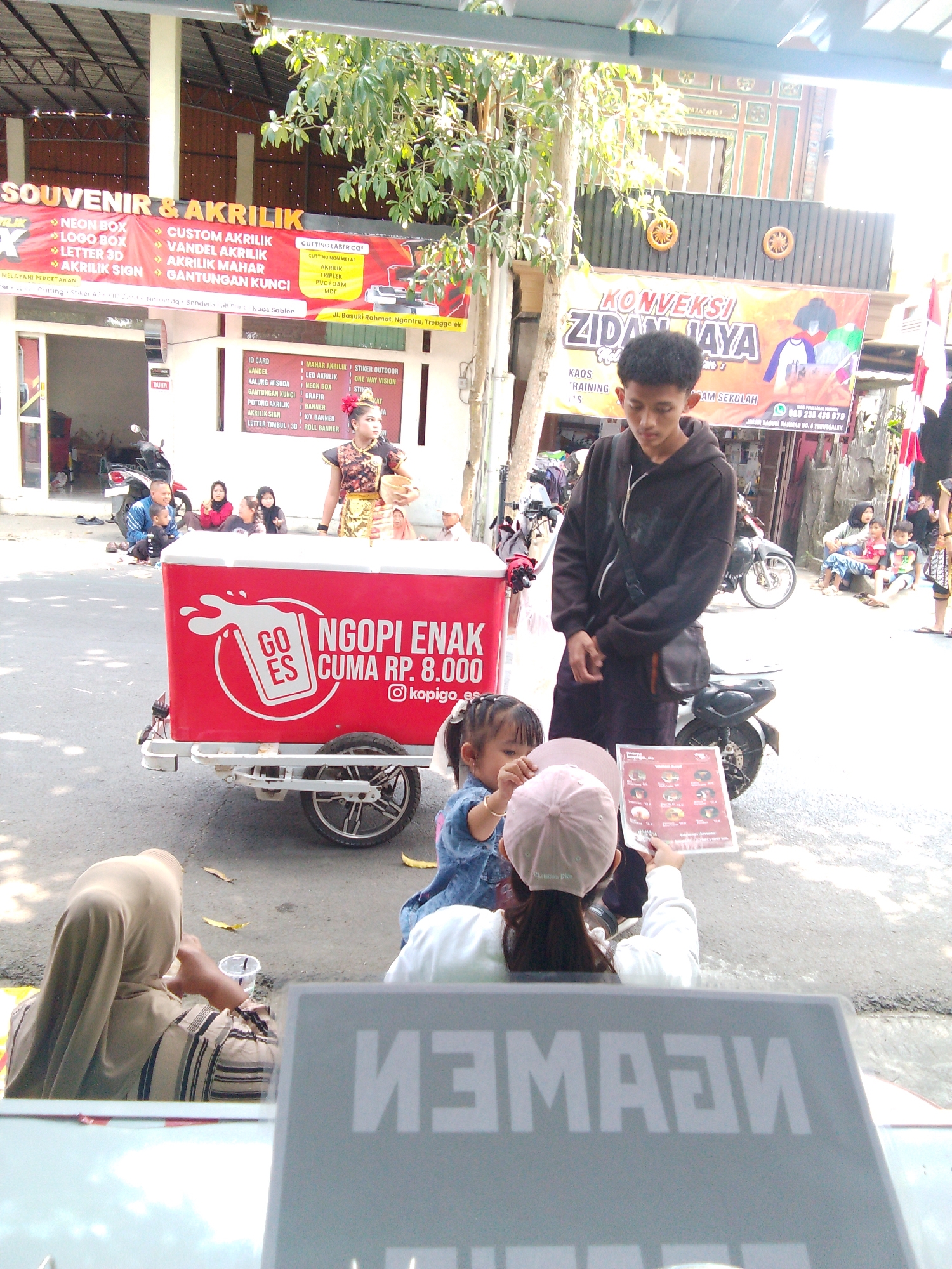 The image depicts an outdoor scene on a street, likely in the city of Trenggalek, Indonesia, as indicated by signage. In the foreground, a red mobile cart with the branding "GO ES NGOPi ENAK CUMA RP. 8.000 @kopigo_es" is prominently displayed, suggesting a beverage or food vendor. A young girl wearing a denim jacket and a pink cap is leaning forward, holding and looking at a menu or pamphlet. To her left, a young man wearing a dark hoodie and a crossbody bag stands looking down. Behind the cart, several people are seated on the ground, including a woman in a hijab and a man with a child. Further in the background, more people are visible sitting or standing. Above the street, banners are displayed, one advertising "SOUVENIR & AKRILIK" with various services listed, and another with "KONVEKSI ZIDAN JAYA". The environment appears to be a commercial or residential street with buildings and trees lining the sides. The overall impression is a bustling street scene with commercial activity and social interaction.
