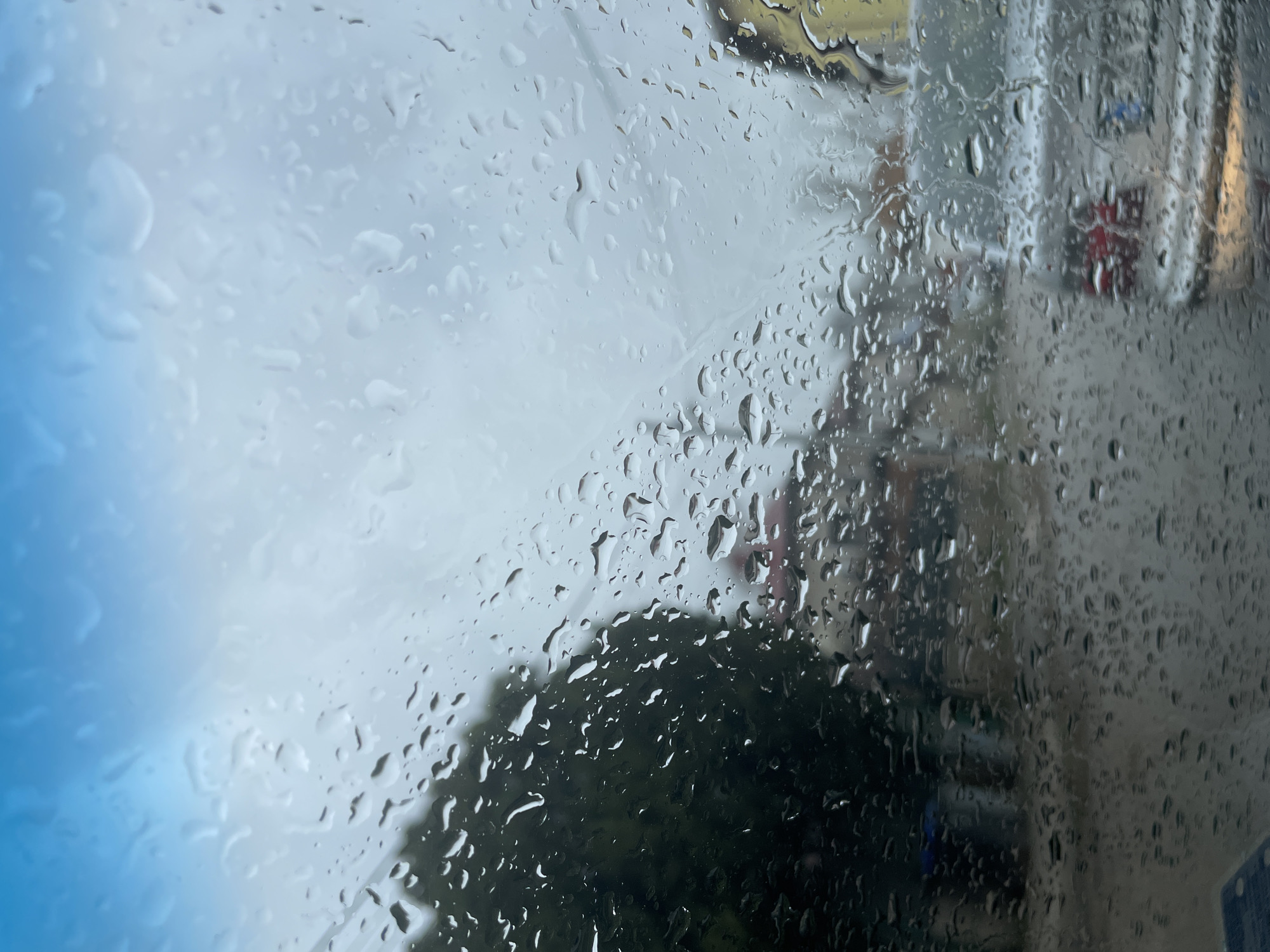 The image shows a view through a rain-streaked window. On the right, there is a blurred view of a building. The street in front is partially visible through the rain, and some trees and bushes are visible. The view on the left shows a sky with cloudy sky. It is impossible to determine the location in Sofia, Bulgaria, based on the provided image.