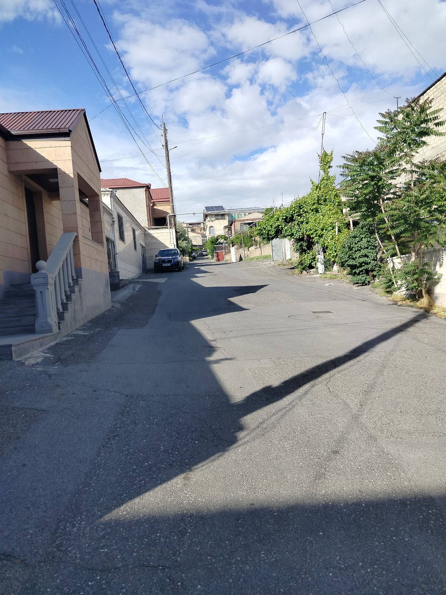 The image depicts a quiet residential street in Yerevan, Armenia. The street is asphalt-paved and slopes slightly uphill. The road is primarily gray and covered in shadow in the foreground. A dark sedan is parked further up the street on the left side. Buildings line both sides of the street, with diverse architectural styles, including a modern building with a brown roof to the left. Vegetation, including trees and vines, is visible on the right side of the street. Utility poles with wires are present above the street. The sky is blue with scattered white clouds. No people are visible in the image.