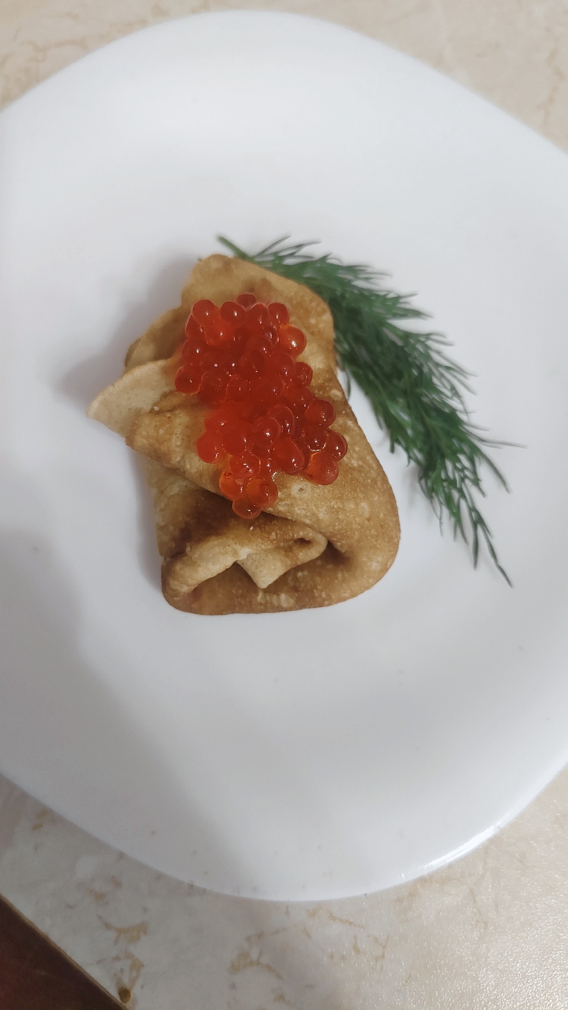 The image displays a close-up of a single serving of a pancake (blin) topped with red caviar and garnished with a sprig of fresh dill. The golden-brown pancake is folded or rolled and occupies the central left portion of a clean, white, round plate. A generous dollop of bright orange-red caviar, with individual translucent roe visible, is piled on top of the pancake. To the right, a vibrant green sprig of dill lies on the plate, serving as a garnish. The white plate rests on a light-colored, possibly marble-patterned, countertop or table surface. The lighting is even, indicating an indoor setting. This presentation is characteristic of traditional Russian cuisine, aligning with the provided location context of Krasnoyarsk, Russia. There is no visible text in the image.
