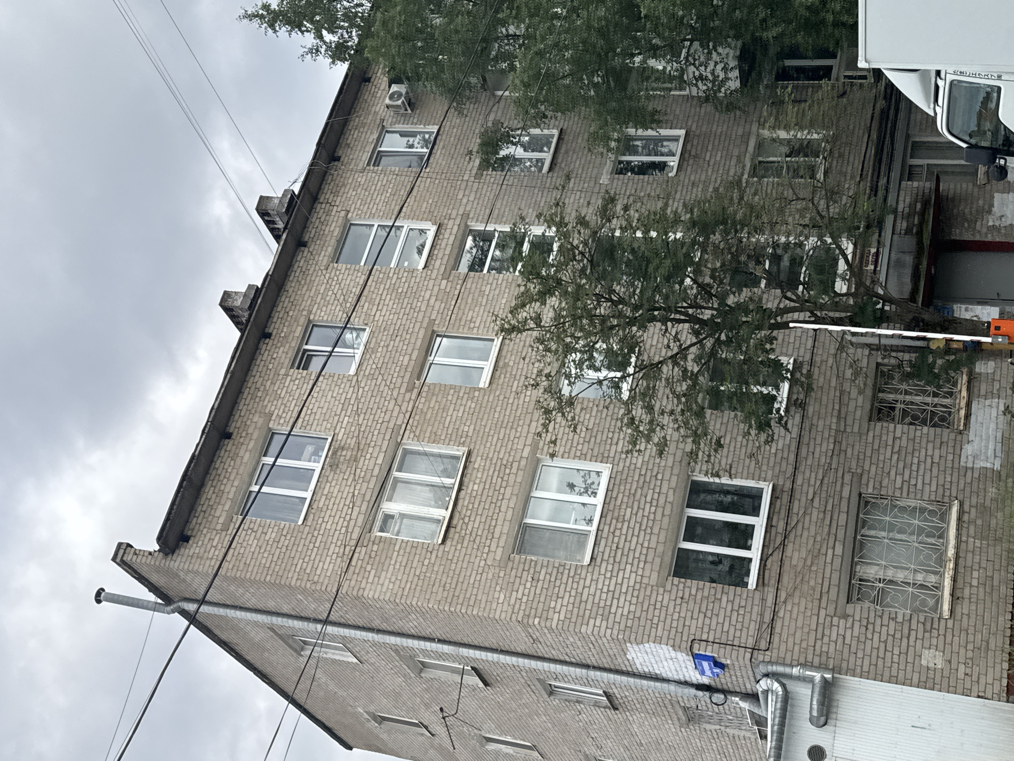The scene depicts a five-story, multi-unit brick building under an overcast sky. The building is constructed of light gray-brown brick and exhibits varying window styles, including modern white PVC frames and older, possibly wooden, frames.
On the left side of the building, a tall, metallic ventilation pipe extends vertically along the corner, culminating in an elbow bend near the roofline. Below it, at ground level, a complex assembly of metallic pipes and a louvered vent is attached to a white, vertical utility structure. A blue street sign, partially obscured by electrical wiring, identifies the location as "ул. Борчанинова" (Borchaninova Street), indicating the city of Perm, Russia. Below the street sign, a patch of white paint is visible on the brickwork. Ground-level windows on this side are secured with elaborate metal security grates.
The roof appears to be flat or low-sloped, featuring concrete coping. Two rectangular brick structures, consistent with chimneys or vents, are positioned on the right side of the roof. Multiple black utility lines traverse the frame diagonally, some attached to the building.
On the building's right facade, an exterior air conditioning unit is mounted on the wall at the third floor from the top. Green-leafed trees partially obscure the lower and middle sections of this facade. At ground level, beneath the trees, an orange and white striped barrier arm is visible. Above a ground-floor entrance, a sign reads "ОДЕЖДА ОБУВЬ" (Clothes Shoes), indicating a retail establishment. The front portion of a white commercial truck, displaying Japanese script on its side, is visible on the far right.
No individuals are visible in the image. The building shows signs of age and weathering, with some visible staining on the brickwork.