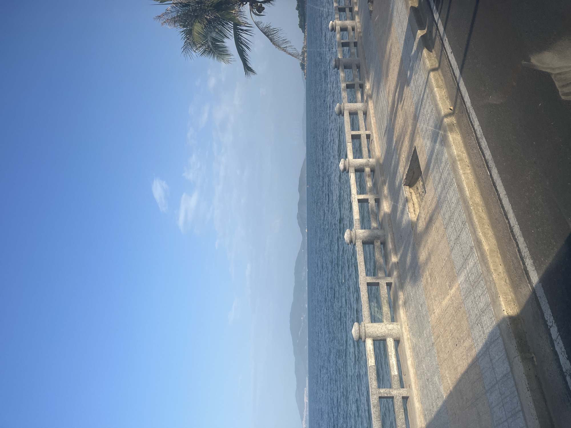 The image shows a view of the sea and coastline in Nha Trang, Vietnam. The foreground features a paved walkway with a stone railing, separated from a road by a concrete curb. The walkway is adorned with patterned tiles in shades of beige and grey, with sunlight casting shadows from the railing. The sea, a deep blue, stretches out to the horizon, where hazy green mountains are visible. A lone palm tree with coconuts is seen on the right side of the frame, partially obscuring the sky. The sky is a clear, bright blue with a few wispy white clouds. The scene appears to be a tranquil coastal promenade.