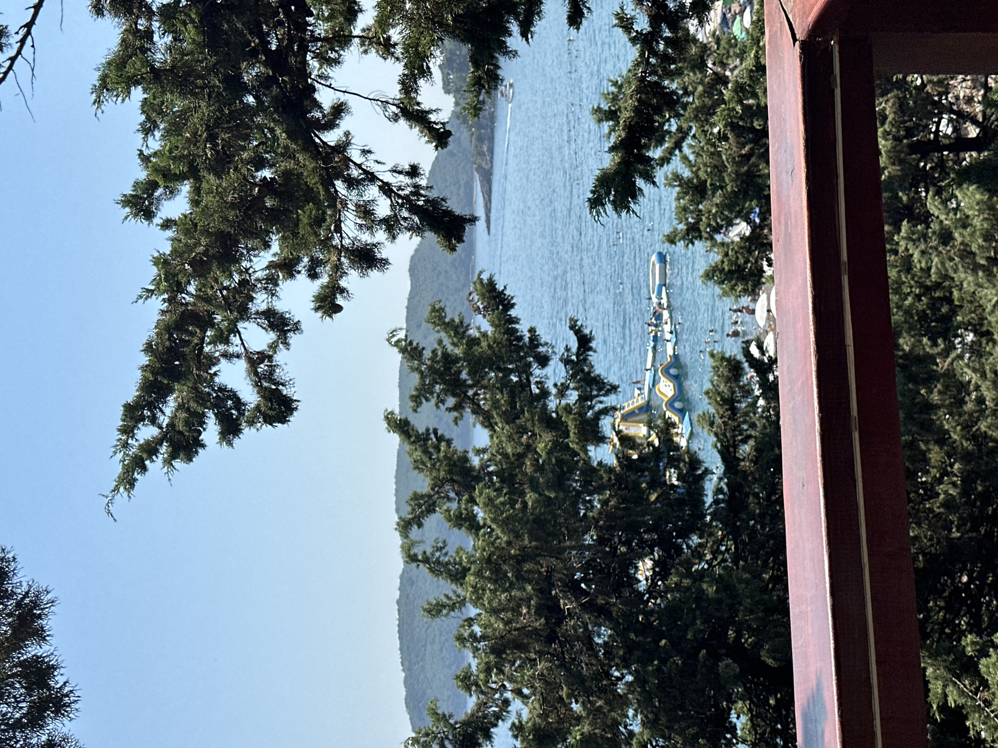 The image depicts a serene coastal scene in Boreti, Montenegro. In the foreground, a dark reddish-brown railing frames the view. Beyond the railing, lush green trees with needle-like leaves partially obscure the scene, creating a natural frame. The middle ground features a sparkling blue sea dotted with several inflatable water toys, including a large yellow and blue structure resembling a water slide or obstacle course. Numerous people are visible in the water, some swimming and others interacting with the inflatable structures. A white boat is navigating the sea in the distance. The background is dominated by rolling, tree-covered hills meeting the horizon under a clear, pale blue sky. The overall impression is one of a sunny day at a popular beach resort.