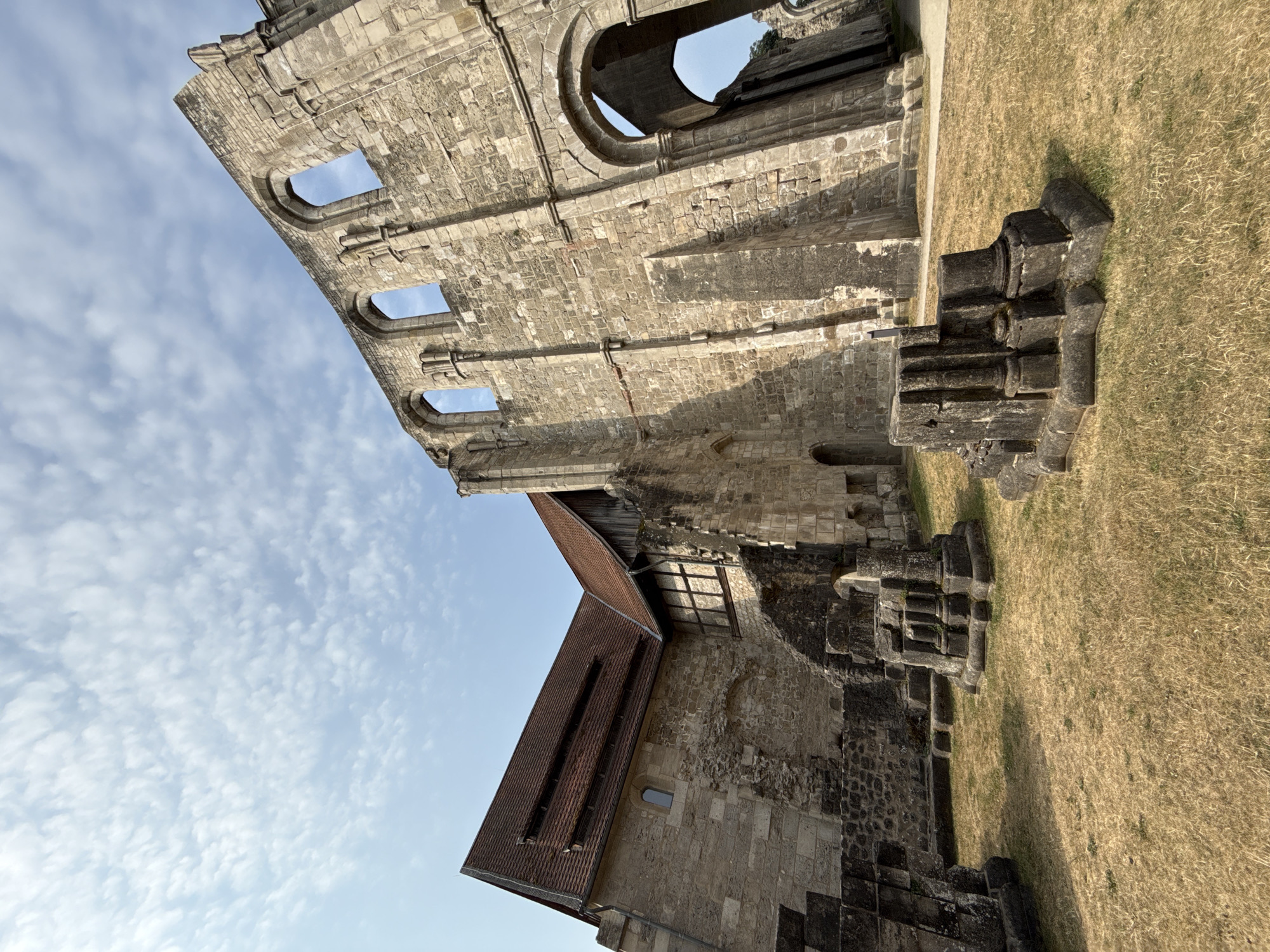 The image depicts the ruins of the Walkenried Abbey in Walkenried, Germany. The foreground features dry, golden grass with several stone fragments scattered across it, resembling carved architectural pieces. In the mid-ground, a well-preserved section of the abbey stands with a red-tiled roof and stone walls, juxtaposed against a large, crumbling stone structure with arched openings that expose the sky. The sky is bright blue with scattered white clouds, suggesting a clear day. The architecture is characterized by massive stone blocks, Romanesque arches, and remnants of Gothic design, indicative of a historical religious site. No people are visible in the frame. The lighting suggests the photograph was taken during daylight hours, with shadows indicating the sun's position.