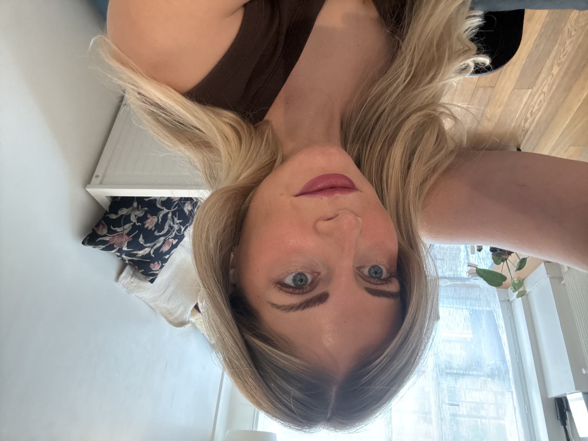 The image features a fair-skinned woman with long, straight blonde hair, parted in the middle, looking directly into the camera. She has blue eyes, prominent eyebrows, and is wearing pink lipstick. She appears to be taking a selfie from a low angle, causing her face to dominate the frame and the overall image to be inverted. She is wearing a dark brown sleeveless top.
When mentally re-oriented, the woman's head is at the top. To her left, a white wall is visible, along with a white wooden shelf. On the shelf, a dark-colored pillow with a floral pattern (pink and green) rests on a light-colored fabric. To her right, a light-colored wooden floor is visible above her shoulder. Below her right shoulder, a window with a sheer, light-colored curtain provides natural illumination to the room. A green plant with broad leaves hangs near the window, and a white radiator or heating unit is visible. The setting suggests an indoor space, likely a home, bathed in natural daylight. There is no visible text in the image. Although the context indicates Paris, France, no specific landmarks or cues from the city are identifiable in the scene.