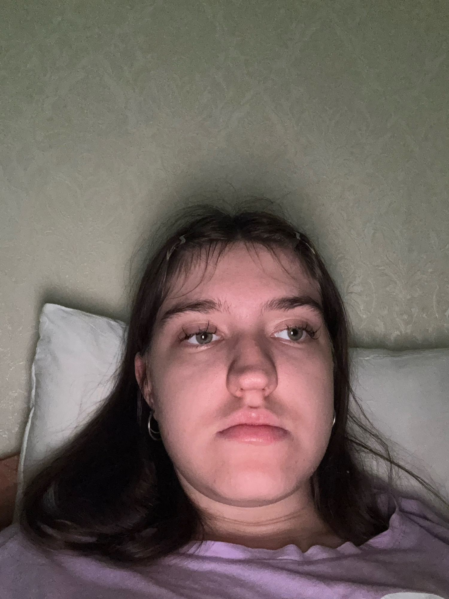 A close-up, head-on, slightly downward-angled selfie of a young Caucasian female with dark brown hair and light green eyes. She is lying down, propped up on a white pillow, with her head against a textured, light green wall. She is wearing a light purple t-shirt with a ruffled neckline. Her facial expression is neutral, with her mouth slightly open. The lighting is dim, suggesting the photo was taken indoors at night. There are no visible landmarks or context to indicate a specific location within Zavolzhye, Russia.