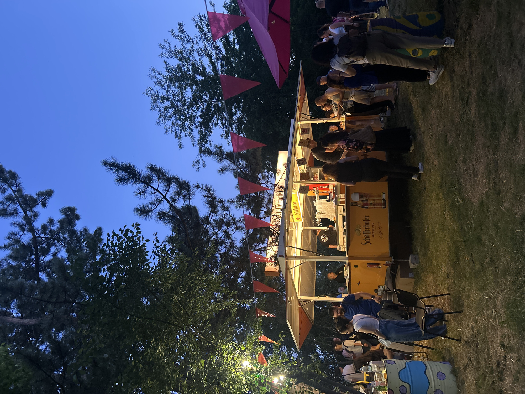 An outdoor scene at dusk features a yellow food truck with the "Schöfferhofer Hefeweizen" logo prominently displayed. The truck has a counter where staff serve customers. Several people are gathered around the food truck, some ordering, others conversing. Red triangular flags are strung above the truck, and string lights illuminate the area. Trees and dark foliage frame the scene, with a clear, dark blue sky above. The ground is covered in grass. A large pink umbrella is visible on the right side of the image, and a decorated barrel-like object is to the left. The atmosphere appears to be that of a casual outdoor gathering or festival.