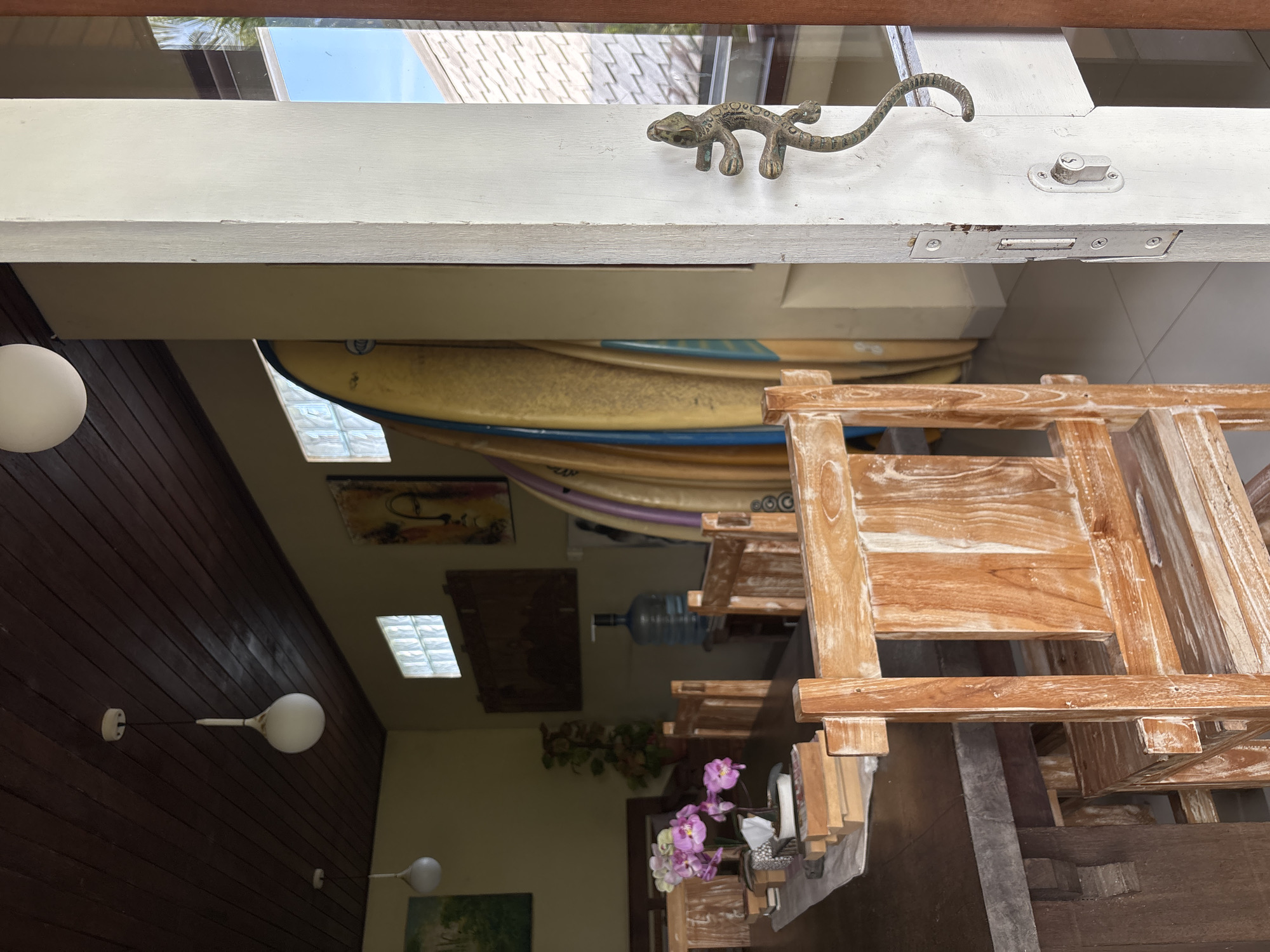 The photograph presents an interior view in Pekutatan, Indonesia, likely of a dwelling or establishment showcasing local decor. A decorative metal lizard adorns a white beam above the main scene. Below, multiple surfboards are stacked along a wall, with one clearly visible. Within the main living space, light fixtures are affixed to a wood-paneled wall. The room also includes art, a water dispenser, and floral arrangements. Furniture is notably crafted from wood, creating a rustic aesthetic. No people are directly visible.