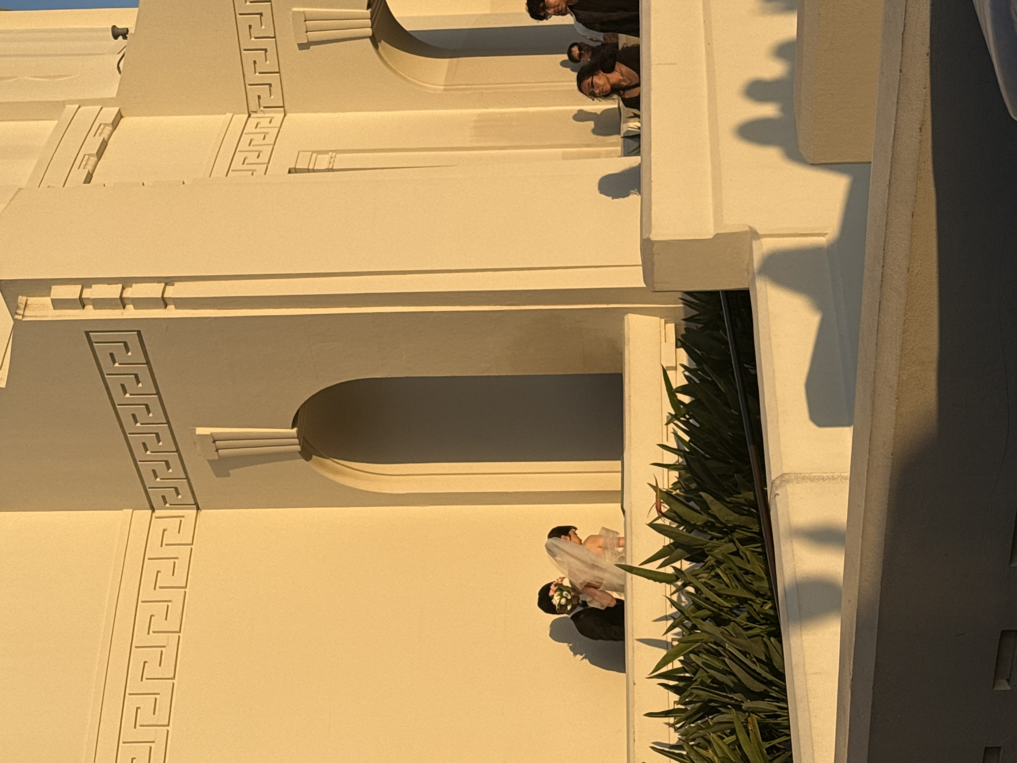 The image shows the exterior of the Griffith Observatory in Los Angeles, California. The building is off-white with a Greek-style design and architectural details. A wedding couple is visible, possibly posing for photos, partially obscured by greenery. Additionally, a small group of people can be seen on a higher level of the building, likely tourists or visitors.